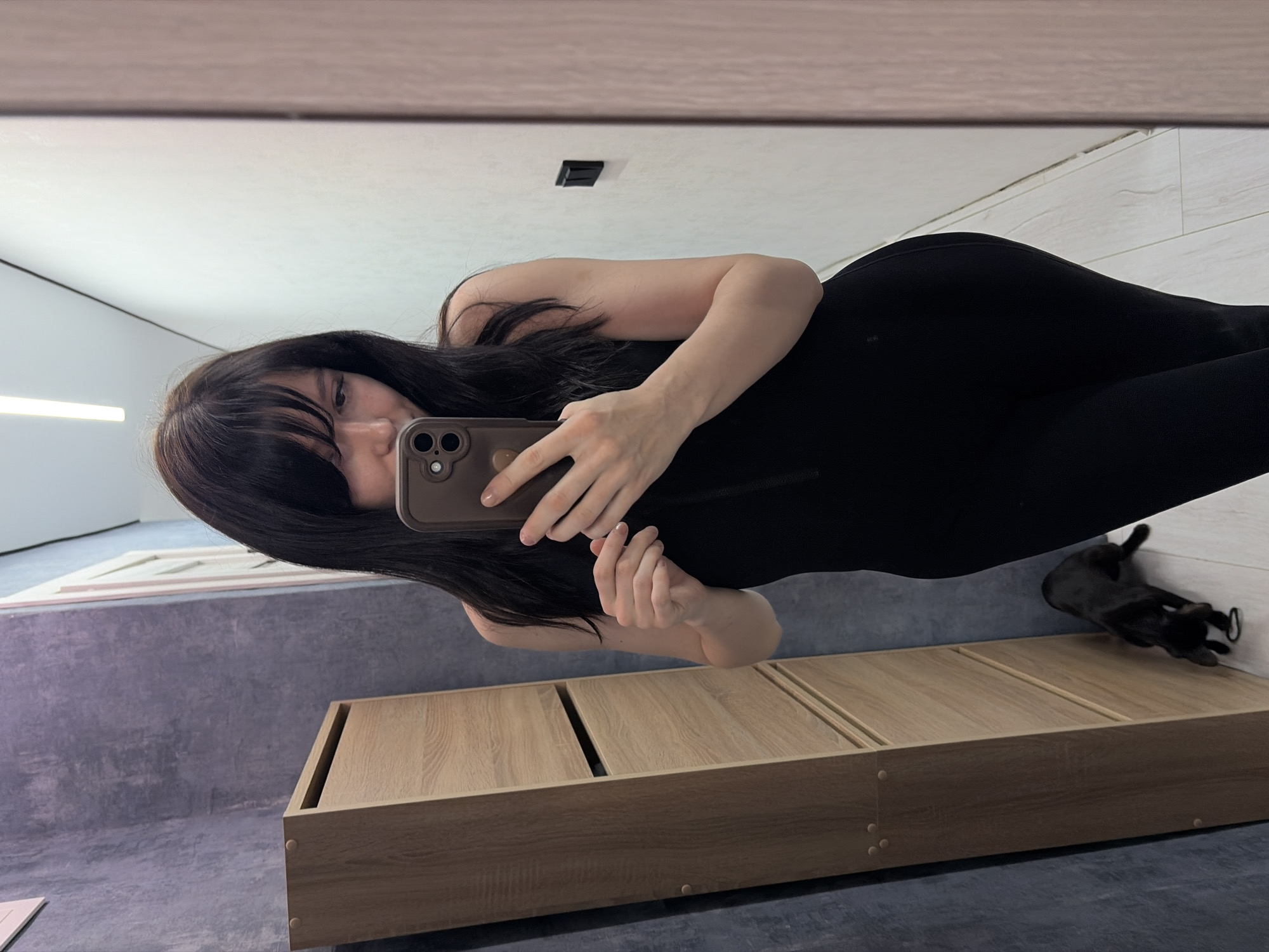 A woman with dark hair and bangs is taking a mirror selfie. She is wearing a black outfit. She is holding a smartphone with a brown case. The background shows a room with a wooden platform and gray walls. A black cat is visible near the wall. The location is Chaikovsky, Russia.