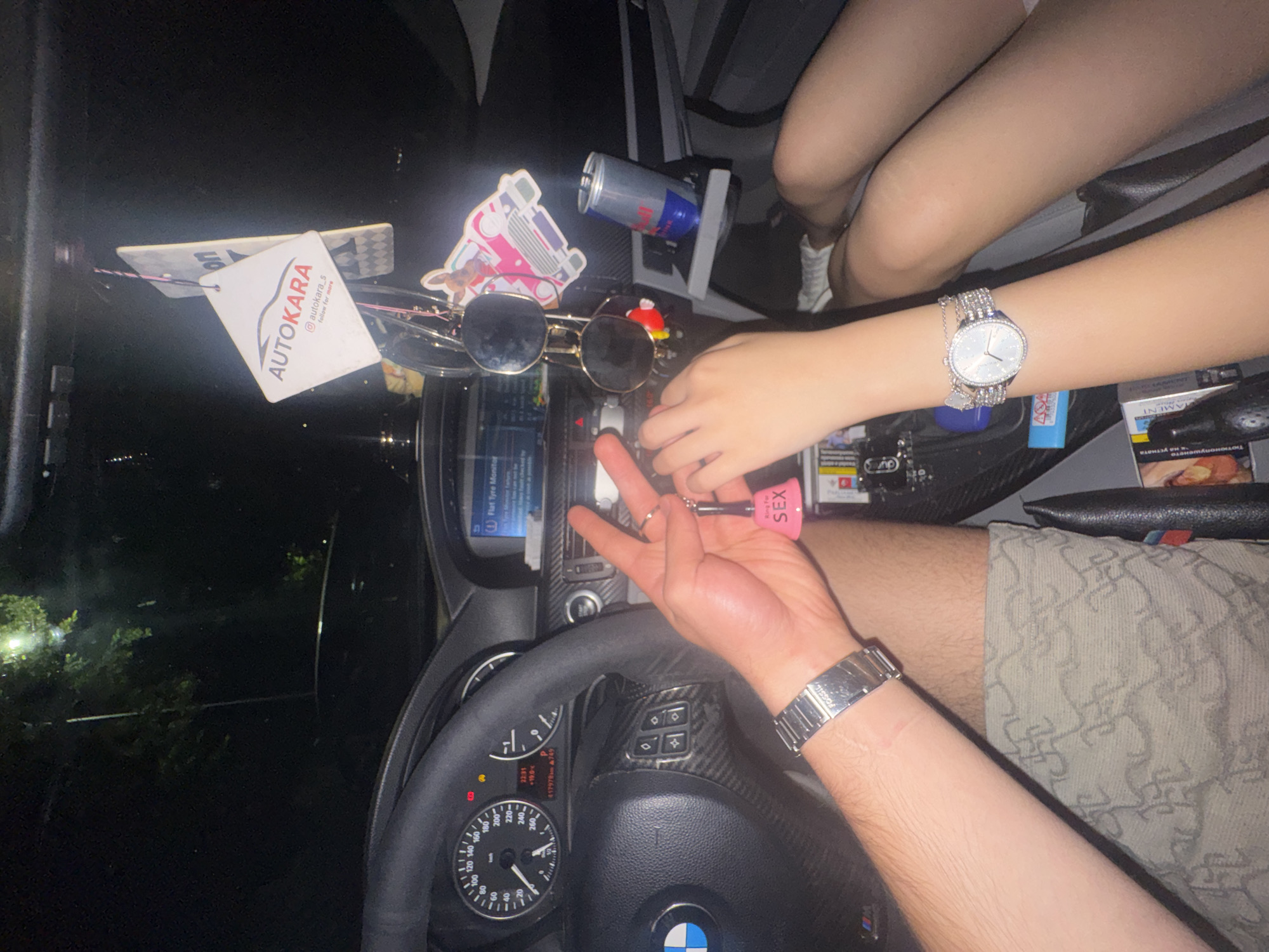 The image captures the interior of a car at night, presenting a casual scene, possibly in Varna, Bulgaria. Two individuals are partially visible through their hands, arms, and legs. One person, with fair skin, has their legs visible, wearing white sneakers. The other individual, whose arm and patterned shorts are seen, wears a silver-toned metallic watch with "FOSSIL" visible on the clasp. This person is holding a small pink keychain that reads "Ring For SEX" and is making a "V" sign with their fingers, while the other person's hand is close by, seemingly interacting with the keychain.
The car's interior features a steering wheel with a BMW logo and carbon fiber trim on the central console. The dashboard instruments are visible, including a speedometer and a digital display. The display shows the time as "22:31", an outside temperature of "+19.0℃", and a mileage of "417978.9". A central screen within the car's system displays "Flat Tyre Monitor".
Various objects are placed on the dashboard and central console. These include a pair of sunglasses, a blue and silver Red Bull can resting in a holder, and a white car air freshener. The air freshener is branded "AUTOKARA" and includes the text "autokara_s follow for more" (likely an Instagram handle). A pack of cigarettes, partially revealing the brand "MAMENT" and some Cyrillic text, is also visible. The person with the fair skin wears a sparkling silver watch. Through the windshield, the dark surroundings with blurry lights and trees indicate the nighttime setting.