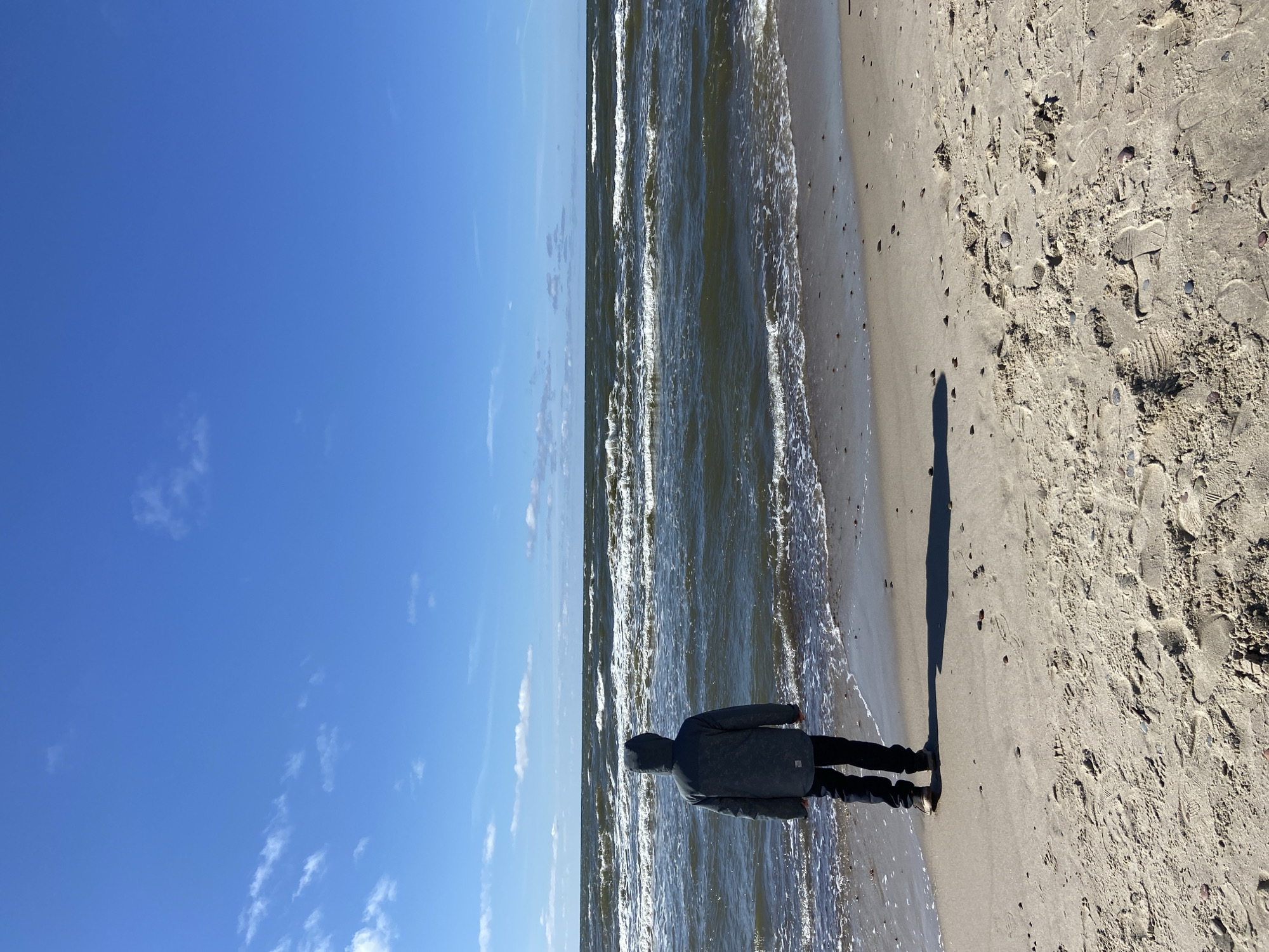 A photograph captures a lone individual standing on a sandy beach at Morskoye, Russia, gazing out at the sea under a bright blue sky. The scene is illuminated by sunlight, indicated by the person's long shadow stretching across the sand. The person is viewed from behind, wearing a dark jacket, possibly hooded, and dark trousers. They stand at the water's edge where gentle waves are breaking on the shore. The sea appears calm with small whitecaps visible on the waves. The beach is composed of light-colored sand, marked with numerous footprints, shells, and small pebbles. The sky above is mostly clear with a few scattered white clouds. The time of day appears to be either early morning or late afternoon, given the extended shadow. The overall mood is peaceful and contemplative. No visible text is present in the image.