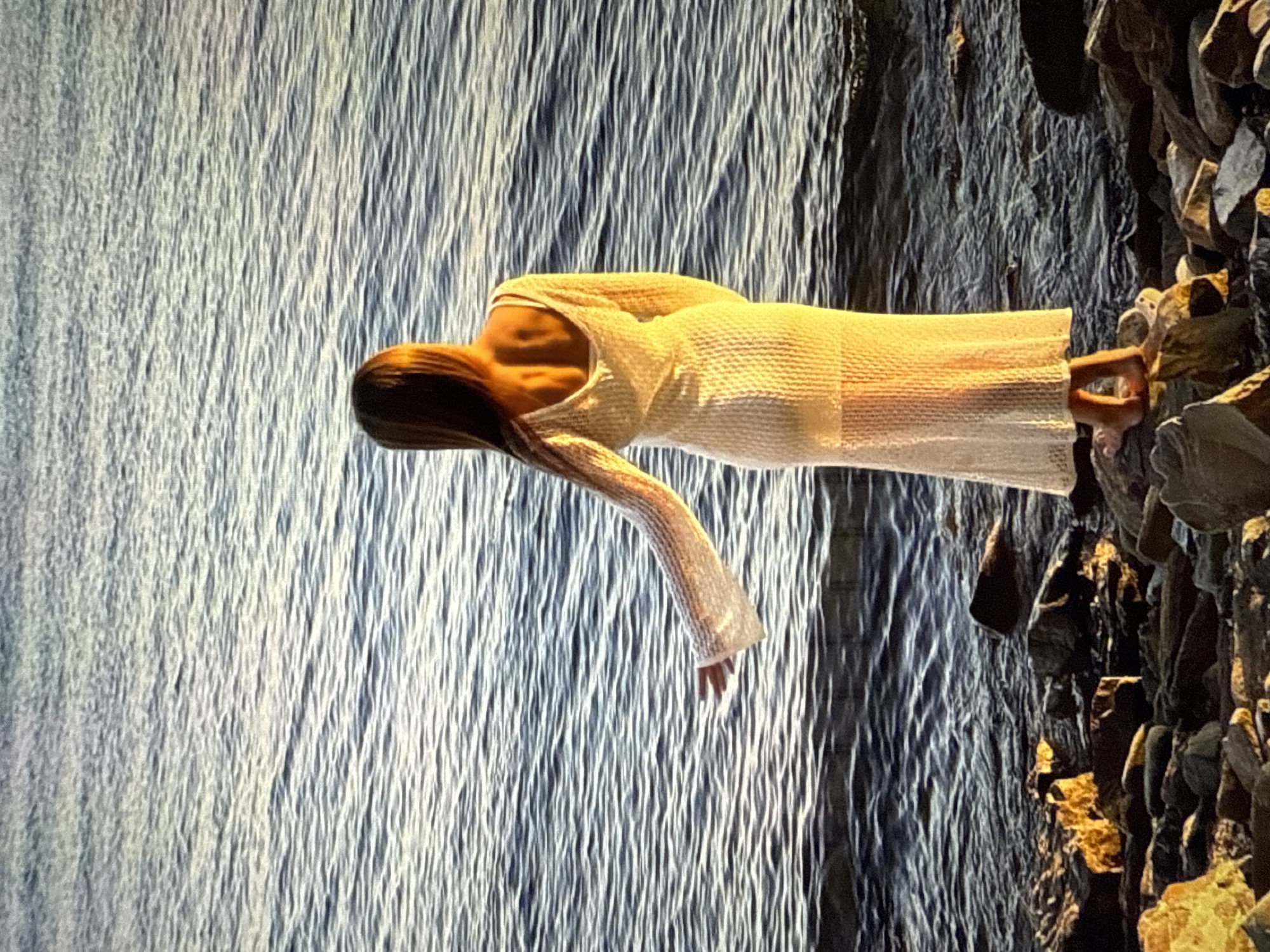 A person wearing a long, cream-colored, knitted dress with long sleeves stands on a rocky shoreline facing the water. The person's back is to the camera, with their hair long and brown. Their left arm is extended to the side, with their hand open. The water is calm with gentle ripples, reflecting the golden light of what appears to be sunset. The shore is covered in dark, angular rocks, some of which are illuminated by the sun. The scene is set in Găvănoasa, Moldova.