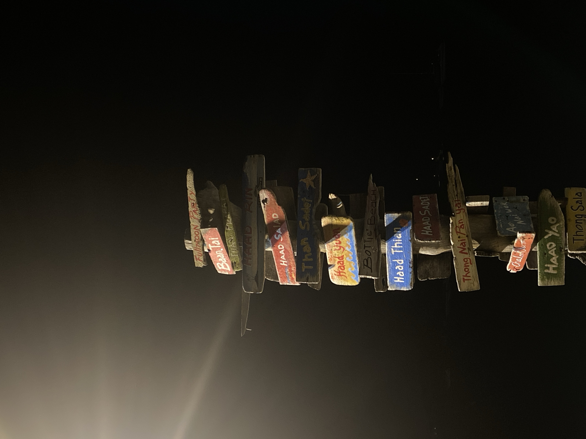 A nighttime view shows a collection of rustic wooden signs displaying place names, presumably destination markers. These signs are suspended from a horizontal support, giving the impression of a directional guide. The signs are painted with various colors, adding a touch of vibrancy against the dark background.
Many place names are visible, including: "Baan Tai," "HAAD RIN," "HAAD SALAD," "Than Sader," "Haad Yoon," "Bottle Beach," "Haad Thian," "HAAD SADET," "Thong Nai Pan yai," "Chaloklum Mai wok," "HAAD YAO," and "Thong Sala."
The scene is dimly lit, with the signs being the primary focus due to a light source illuminating them. The overall atmosphere suggests a tourist-oriented location, possibly in a town or near popular beaches. The location is Baan Bang Rak, Thailand.