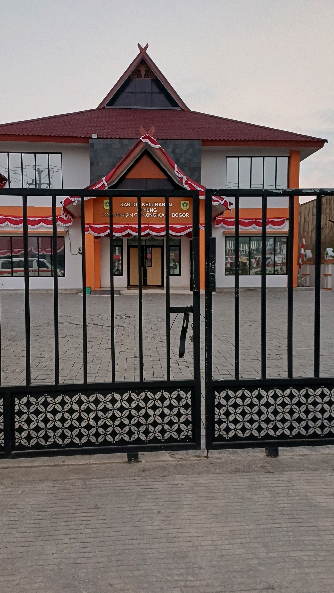 The image shows the exterior of a building identified by the text "KANTOR KELURAHAN" and "KECAMATAN CIBINONG KAB. BOGOR". The building has an orange and white facade with a dark red tiled roof. The entrance is framed by a triangular awning decorated with red and white bunting. A black metal gate with a decorative pattern is in the foreground, partially obscuring the view of the building and a paved courtyard. The sky above is overcast. There are no people visible in the image.