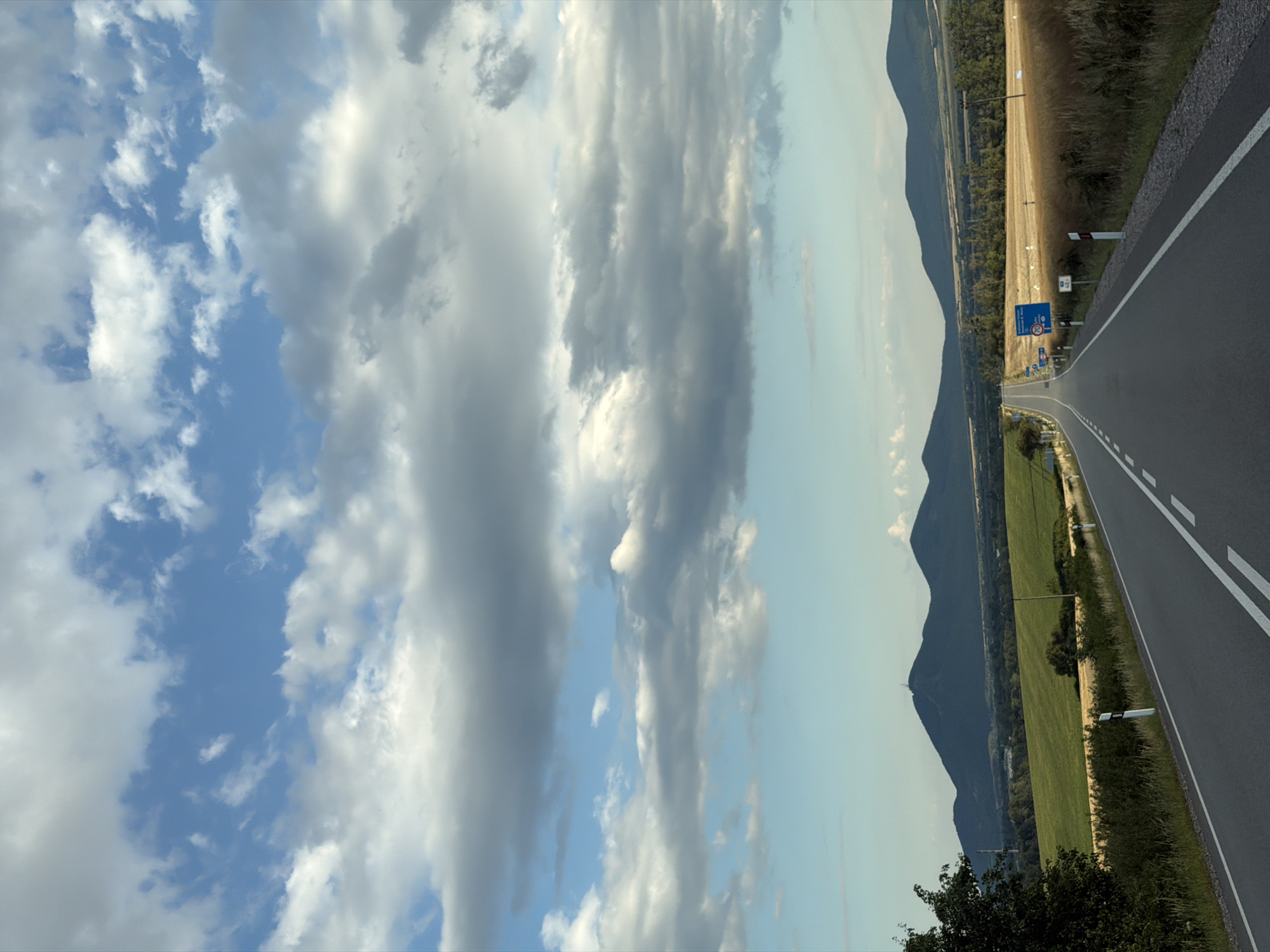 The image depicts a landscape view from a road in Luhyňa, Slovakia. A paved road with white lane markings extends into the distance, flanked by fields of green and golden crops. Rolling hills and dark, conical mountains form the backdrop under a sky filled with scattered white and gray clouds. Several blue road signs are visible along the side of the road. The scene appears to be an outdoor, rural environment during daylight. There are no people or animals visible in the image.