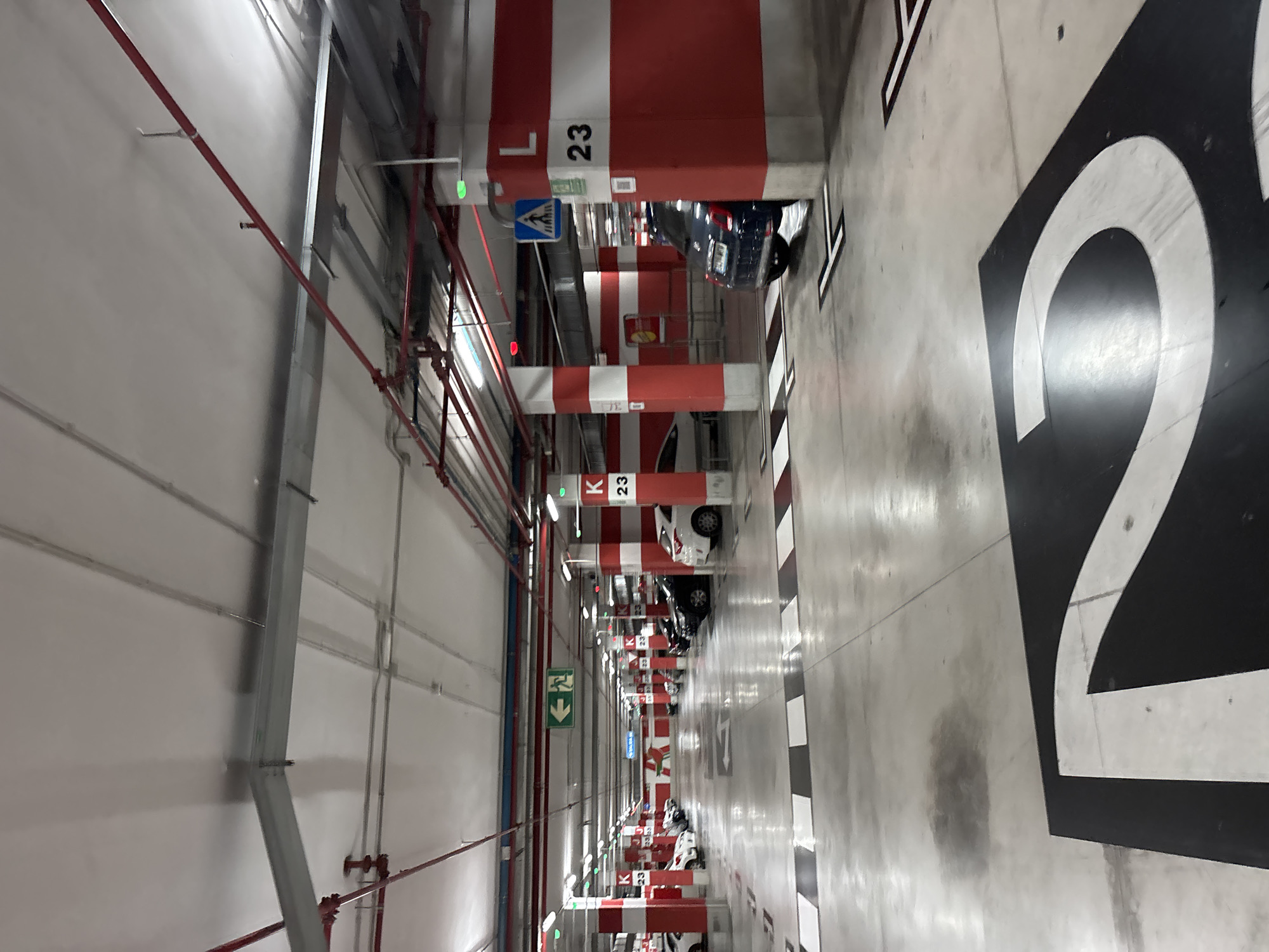 The image depicts an indoor parking garage, likely located in Orihuela, Spain, as indicated by context. The environment is characterized by concrete flooring and ceilings, with numerous white and red striped pillars supporting the structure. Overhead, an array of pipes and lighting fixtures are visible. The floor is marked with directional arrows and black and white striped pedestrian crossings. Various cars are parked in designated spots, with some visible in the background. Signs indicating parking zone letters and numbers are present on the pillars, such as "K 23" and "L 23". A pedestrian crossing sign with a blue background and a white silhouette of a person walking is also visible. The overall lighting suggests an artificial environment. The image is captured from a low to medium angle, looking down a lane of the parking facility.