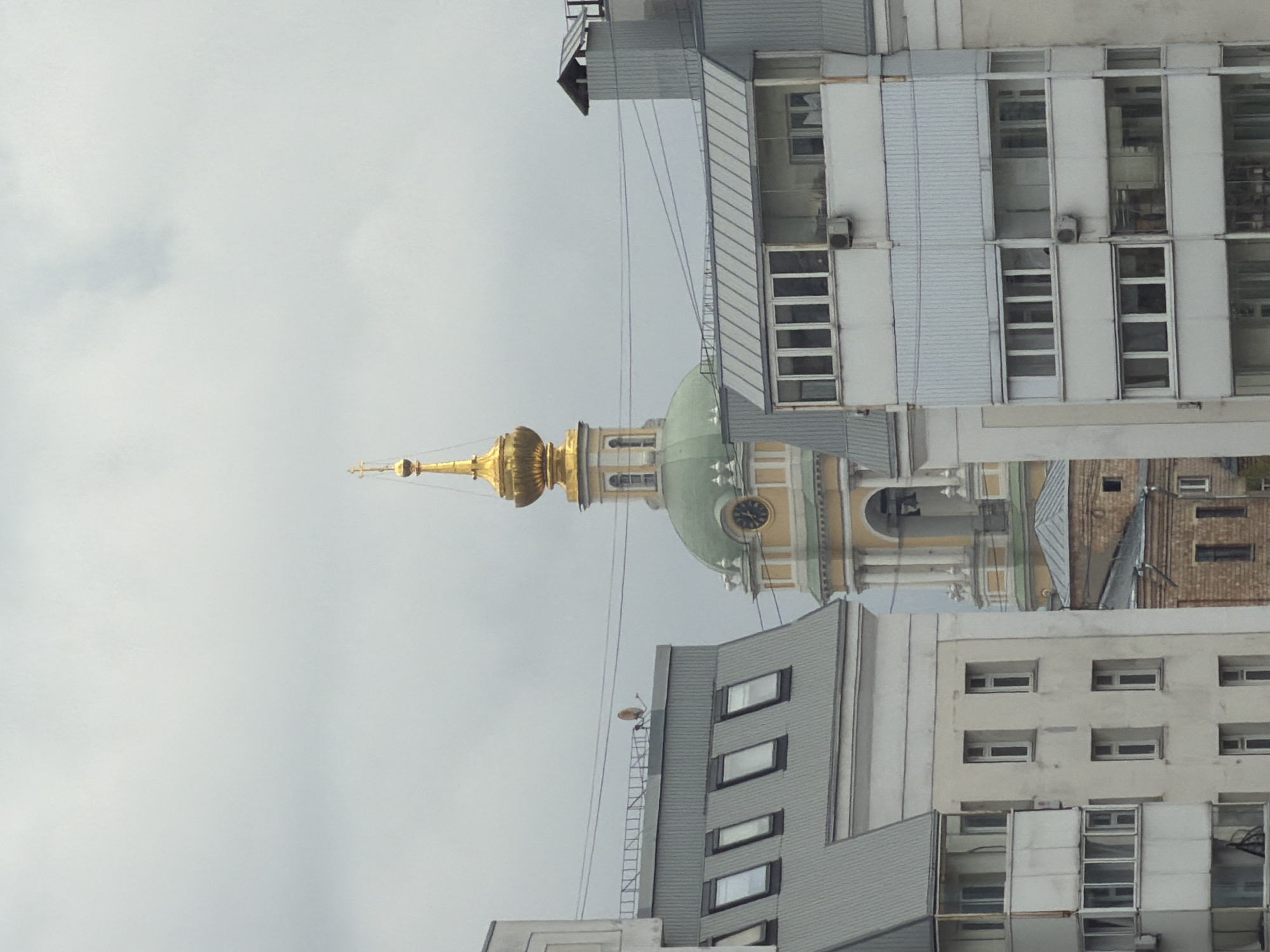 The image displays an urban landscape in Moscow, Russia, captured under an overcast sky. The photograph is oriented vertically, showcasing a juxtaposition of historical religious architecture and contemporary buildings.
At the central part of the frame stands a grand, golden-domed church or cathedral, topped with a golden spire and a cross. Below the gleaming golden dome, a smaller green dome with a visible clock face is present, followed by classical architectural elements such as columns and arches in shades of light yellow and white.
Flanking and surrounding this historical structure are several multi-story modern buildings. On the upper right, a large building features a grey, possibly metallic, corrugated facade with numerous rectangular windows and visible air conditioning units. In the lower right foreground, another building with white walls displays rows of smaller, uniformly spaced windows, some with white frames. Another grey building with larger, modern windows is visible on the bottom left. Thin power lines or communication cables traverse the middle ground horizontally, connecting different structures. A small satellite dish or antenna is visible on the rooftop of one of the grey buildings.
The sky is a uniform light grey, indicating an overcast or cloudy daytime. No people or specific events are depicted, and there is no visible text. The scene presents a blend of the city's rich historical heritage and its ongoing urban development.