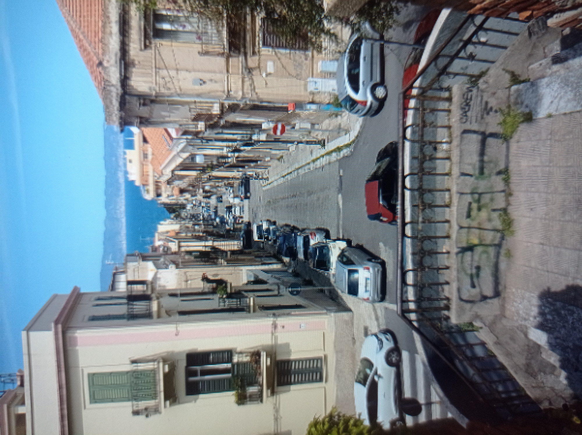 The image shows a narrow, steep street lined with multi-story buildings and parked cars, with the sea visible in the distance. The buildings have balconies and windows, and some have potted plants. The street is paved and appears to be a typical European urban scene. There are several cars parked on both sides of the street, and some are in motion further up the hill. The sky is clear and blue. The foreground shows a concrete staircase with a metal railing, with some graffiti visible on the steps. The location is not in Mukachevo, Ukraine; it is likely a coastal town in Italy given the architecture and the presence of the sea.