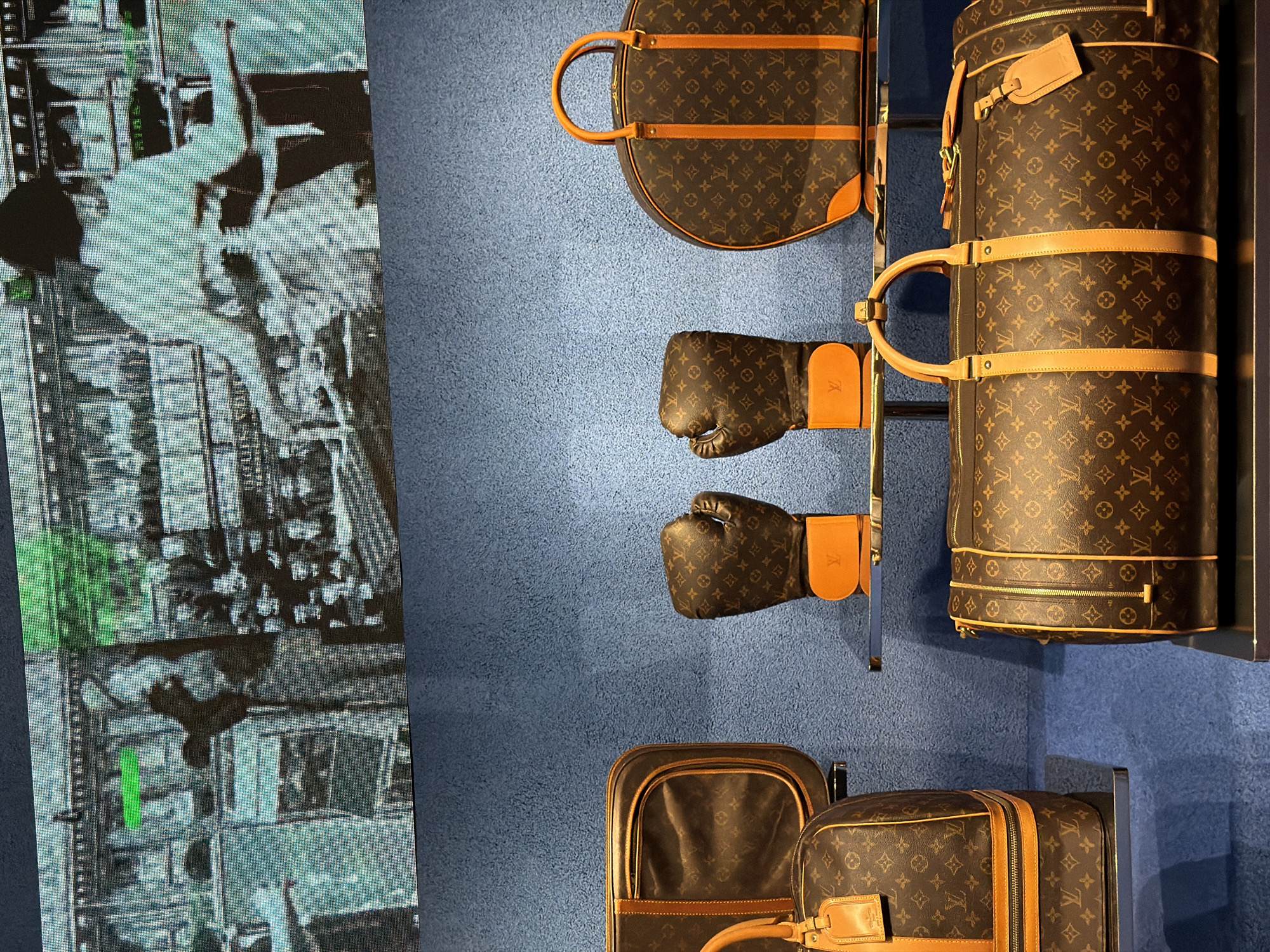 The image shows a display of Louis Vuitton luggage and boxing gloves in front of a screen showing a black and white image of people in motion. The luggage and boxing gloves are in Louis Vuitton's signature brown monogram pattern with tan leather trim. The items are arranged on a blue wall. The black and white image on the screen is difficult to discern, but appears to show people interacting or moving.
While I don't have information to precisely pinpoint the exact location within Jing'an District, China, it's likely this display is in a high-end retail environment, such as a department store or a Louis Vuitton boutique.