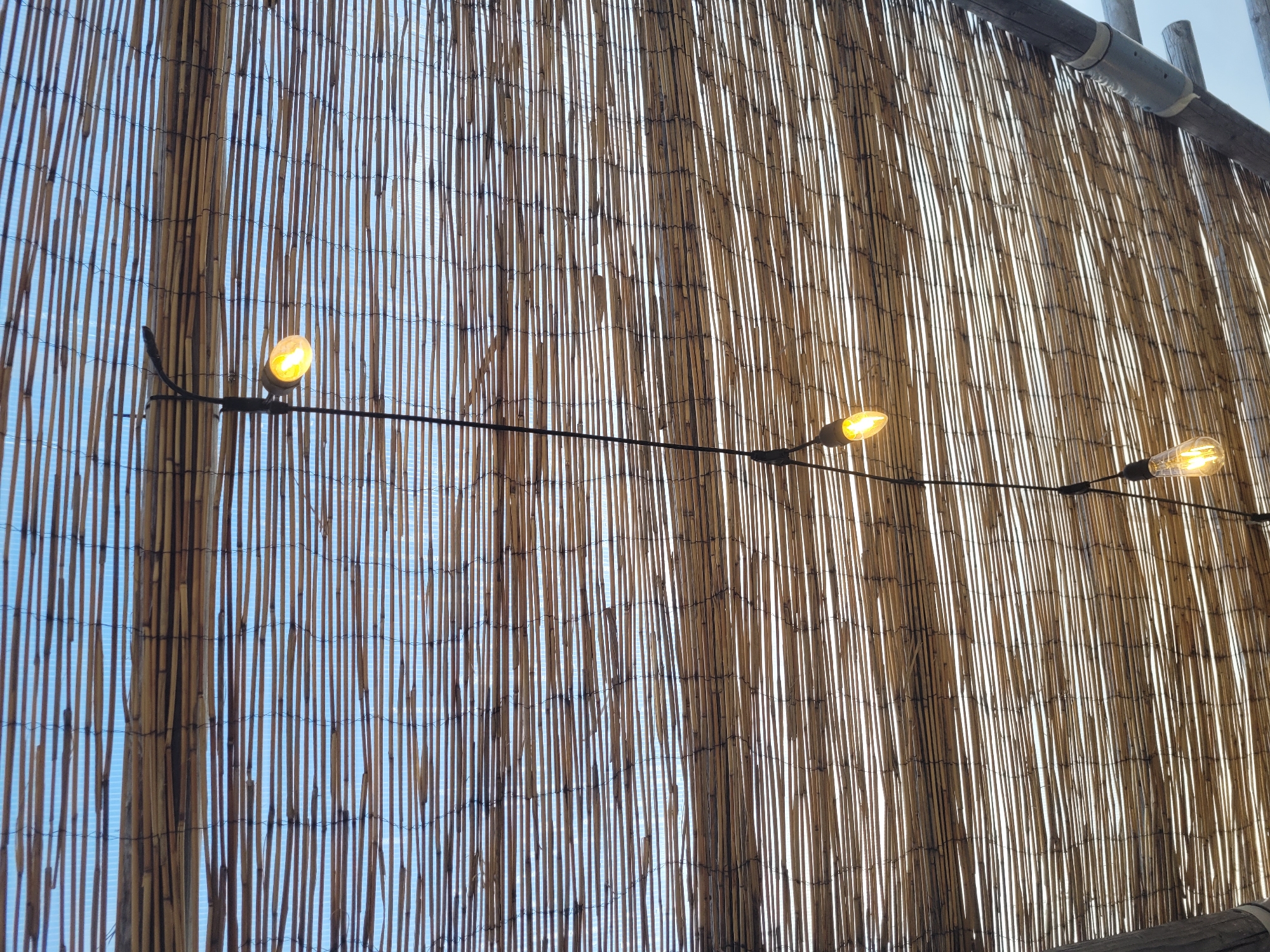 An outdoor scene in Jūrmala, Latvia, features a rustic bamboo awning stretched across a wooden frame. Suspended from a black cord, a string of four vintage-style Edison bulbs illuminates the underside of the awning. The bulbs, varying in their illumination, cast a warm glow through the gaps in the bamboo. The sky is visible through the awning, appearing bright and possibly overcast or hazy. The overall impression is one of a relaxed, possibly seaside, setting.