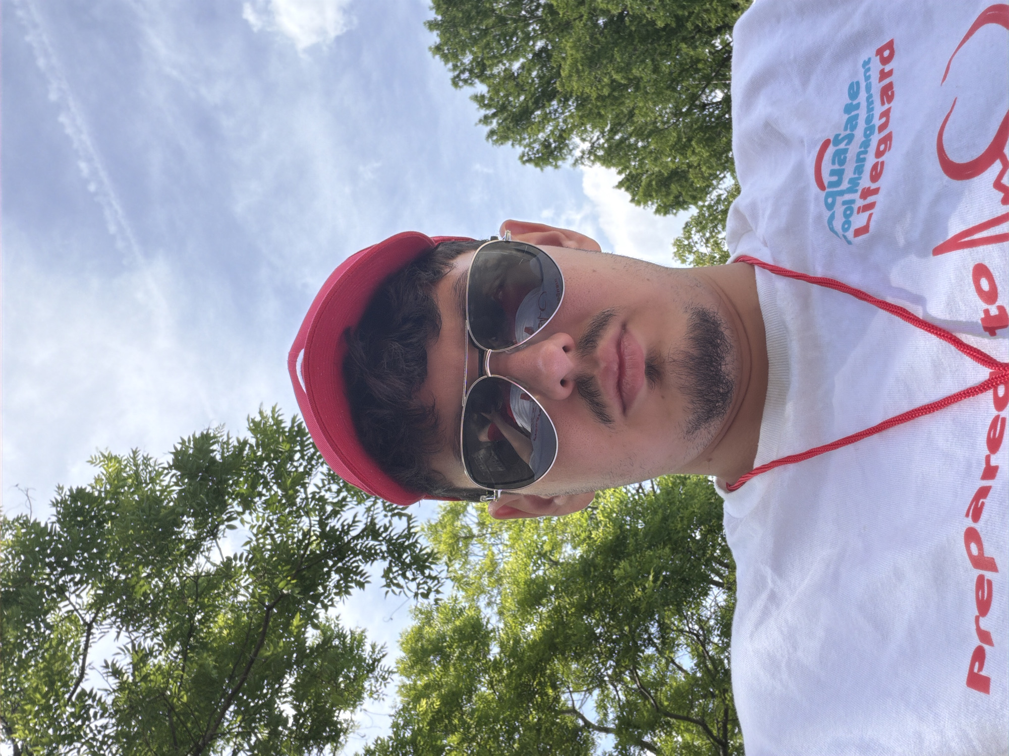 A man wearing a white shirt with the Aquasafe Pool Management Lifeguard logo and the word "Prepared" is shown in a selfie taken outdoors, possibly in Edison, United States of America. He is also wearing a red hat and sunglasses. Trees and a partly cloudy sky are visible in the background.