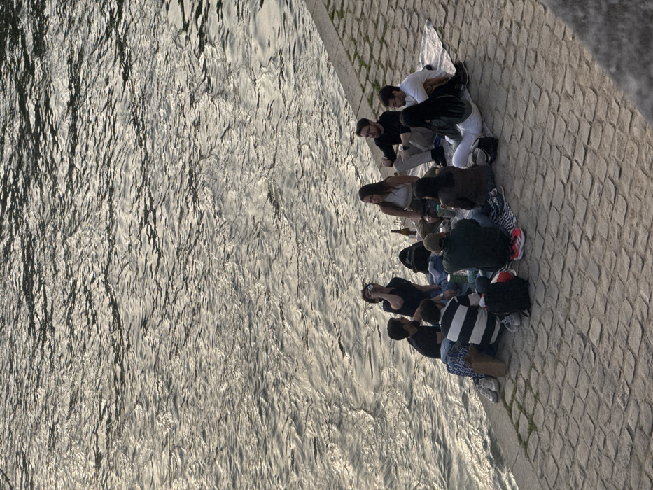 Ten young adults are seated in a semi-circle on a paved riverbank next to a body of water, likely a river, in the city of Unknown, Unknown. The water exhibits ripples and reflects ambient light. The group sits on various light-colored and striped blankets or garments spread across reddish-brown cobblestones. Individuals are dressed in casual attire, including t-shirts, a tank top, a striped long-sleeve shirt, and jeans or casual pants. One individual on the left side is actively drinking from a silver can. Other objects present include a brown glass bottle, additional cans, a black backpack, a brown paper bag, and various pairs of shoes placed around the perimeter of the seating area. The individuals appear to be engaged in social interaction.