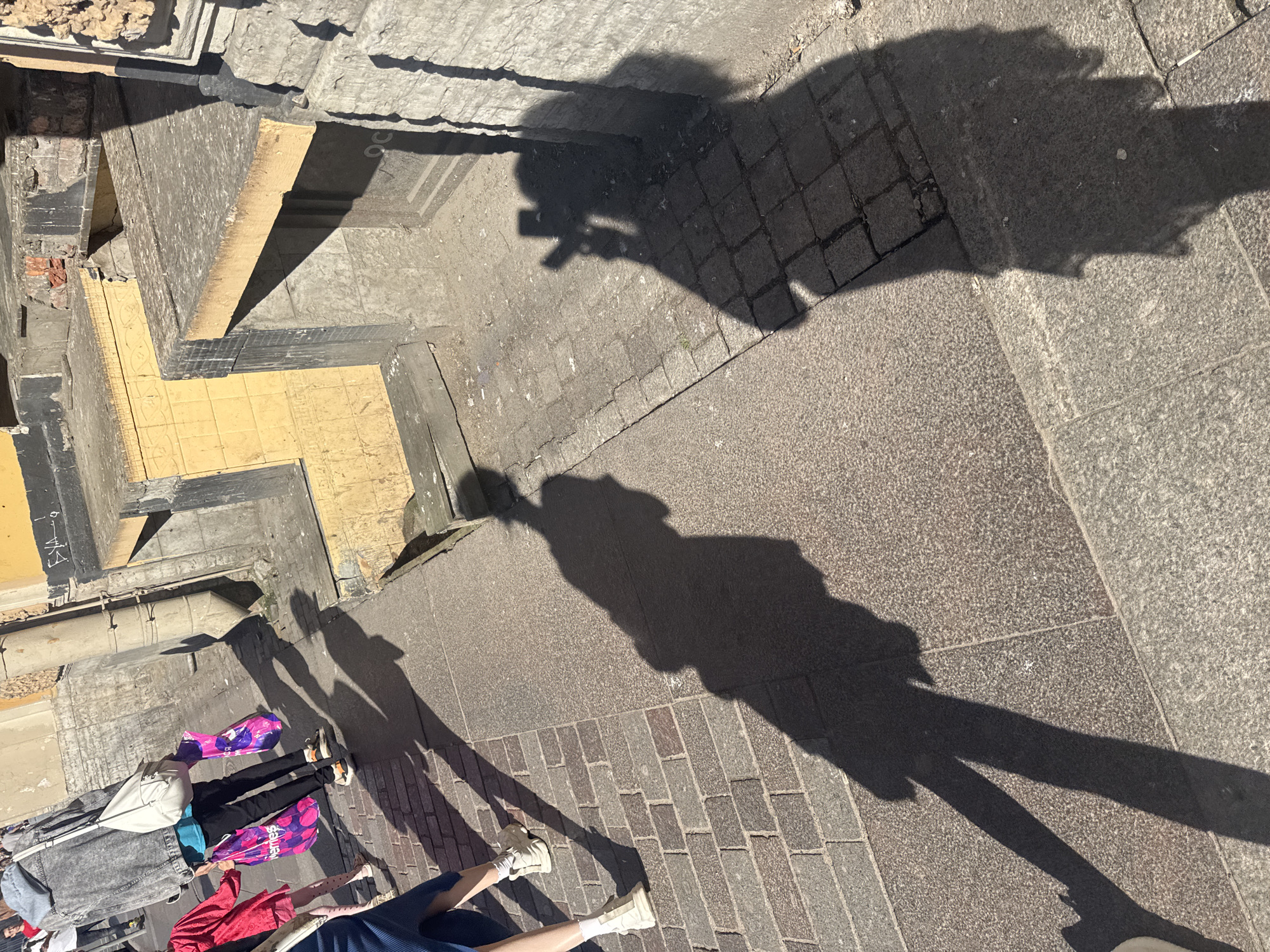 The image captures a sunlit street scene in Saint Petersburg, Russia. The foreground displays two distinct shadows on a cobblestone and granite pavement. The shadow on the left depicts a person with their legs extended, wearing white socks and light-colored shoes. To the right, a larger shadow shows a person holding a smartphone, appearing to take a photograph. The background reveals a building with a yellow tiled facade and weathered brickwork. A vertical pipe runs down the side of the building. Further to the left, fragments of other pedestrians are visible, including someone in a red jacket holding shopping bags, and another person in a grey denim jacket carrying a white bag. The overall impression is a candid street view on a bright day.