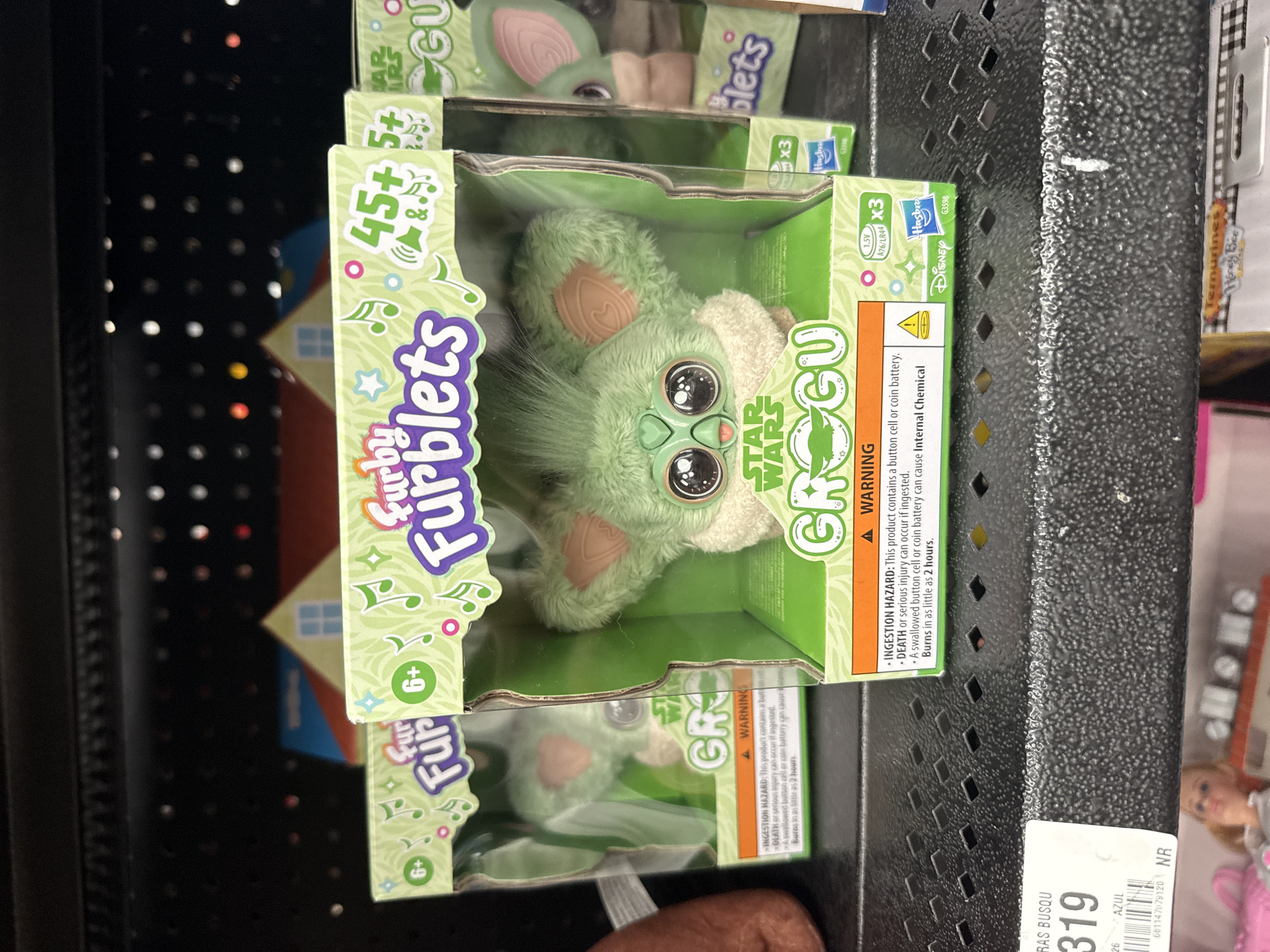 The image shows a close-up of a shelf stocked with "Furby Furblets Star Wars Grogu" toys. The toys are in their original packaging, featuring images of the Grogu character (often referred to as "Baby Yoda" from "The Mandalorian" series) and branding from "Star Wars", "Disney", and "Hasbro". The packaging includes safety warnings regarding ingestion hazards due to the button cell or coin battery. The toys are stacked on a dark metal shelf, with other items partially visible in the background, including a price tag with "319" visible and a doll. The location is San Bernardino Tlaxcalancingo, Mexico is not evident from the image alone.