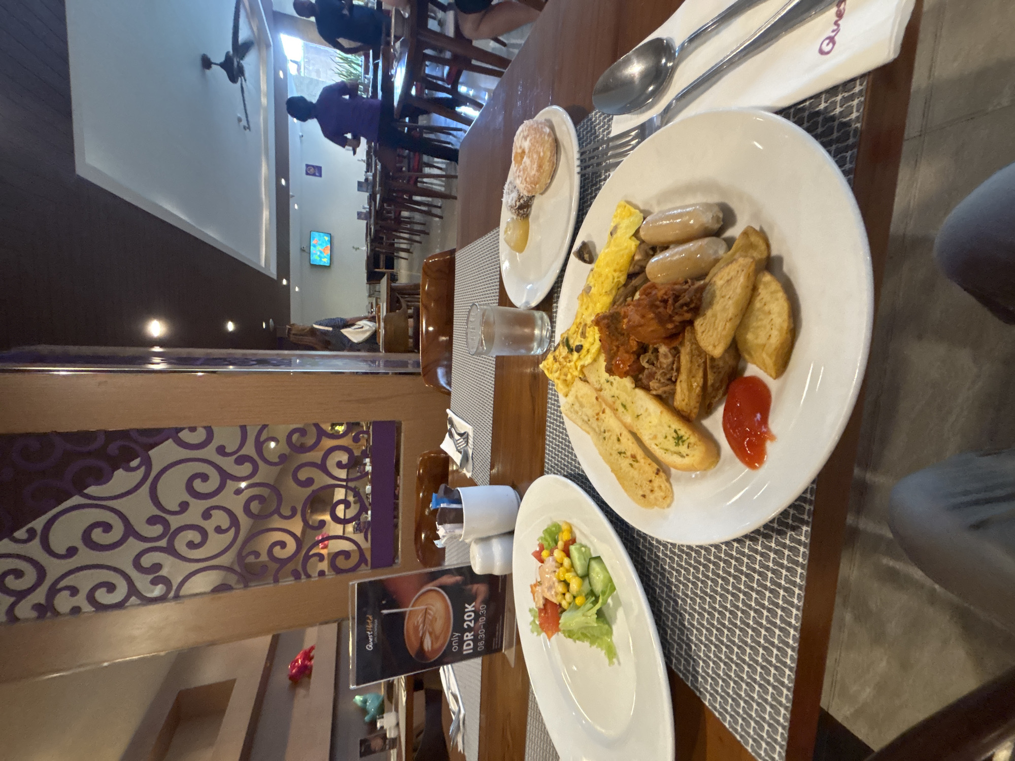 This image depicts a breakfast buffet at the Quest Hotel Denpasar, Indonesia. The foreground features two plates of food. The plate on the left contains a salad with lettuce, corn, tomatoes, and a dressing. The plate on the right displays a variety of breakfast items including scrambled eggs, sausages, fried potatoes, and what appears to be shredded meat or pulled pork, along with a dollop of ketchup. A glass of water and a small plate with donuts and a pastry are also visible on the table. The background shows a dining area with tables and chairs, and several people are present, including hotel staff. The interior design features wooden elements and purple decorative patterns. A promotional stand for the hotel's breakfast offer is also in view.