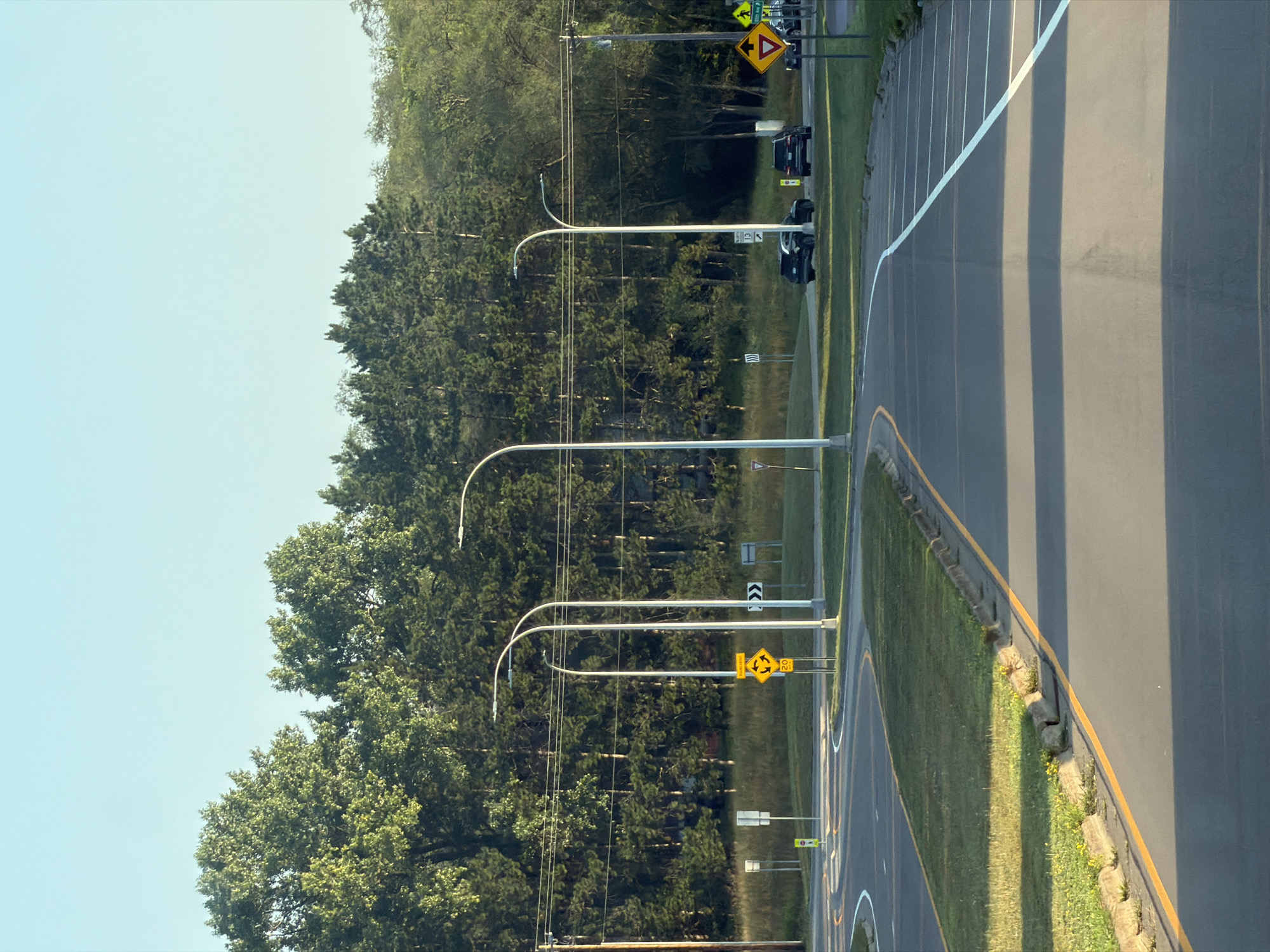 The image shows a road intersection in Elk River, United States of America, under a clear, bright sky. The foreground is dominated by an asphalt road with clear lane markings. A bright yellow line runs along the right edge of the road, separating it from a grassy area with a concrete curb. In the mid-ground, several tall streetlights with curved tops are visible. Road signs are scattered throughout the scene, including a yellow diamond-shaped sign with a circular arrow indicating a roundabout, a speed limit sign reading "20," and a yield sign. A few cars, including two SUVs and a sedan, are present on the road in the mid-ground. The background features a dense line of green trees.