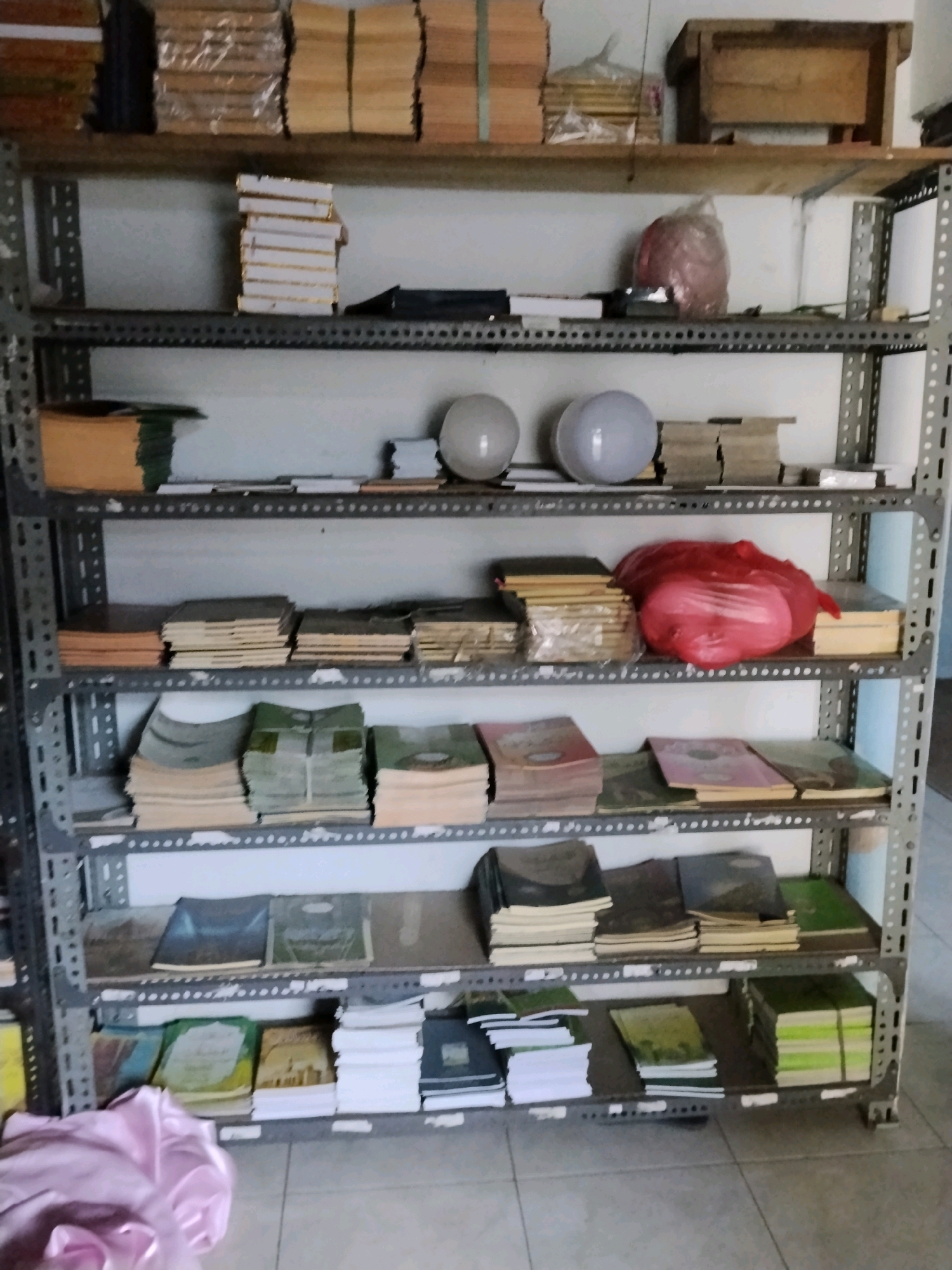 This image shows a metal shelving unit filled with stacks of books and other assorted items. The shelves are organized and appear to be in a storage area or a room with an unfinished ceiling. The items on the shelves include various types of books, some in bundles and others loose. There are also some round objects that resemble light bulbs or decorative spheres on one shelf, and a large red plastic bag containing what appears to be fabric on another. The overall environment suggests a place where books are stored, possibly a library, archive, or a personal collection. The presence of a wooden box on the top shelf and the general arrangement of items do not offer specific clues about the location within the city of Kejawan, Indonesia, beyond it being an indoor storage space.