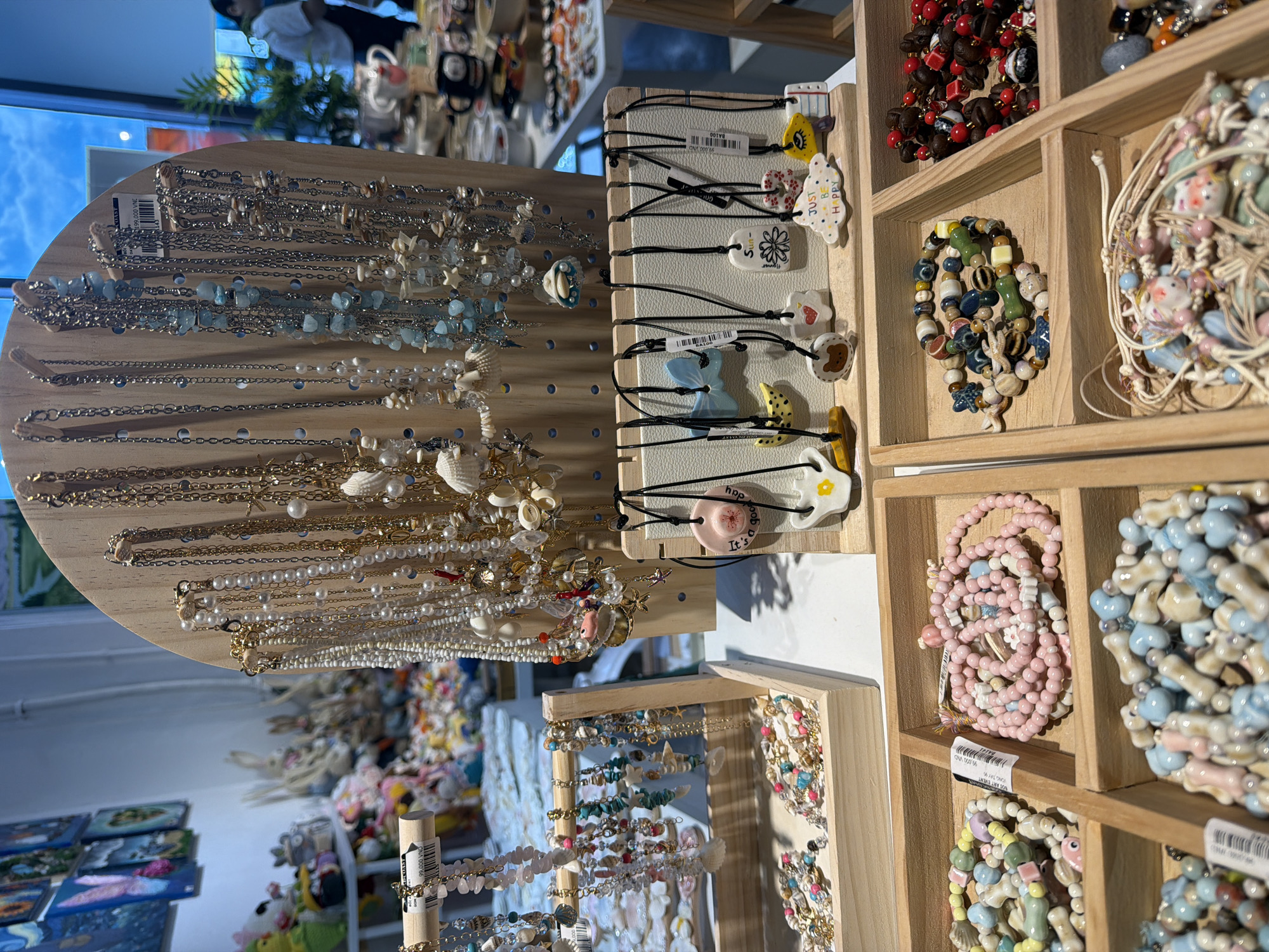The image shows a retail display of various handmade jewelry, including necklaces and bracelets, in a store. The jewelry is primarily made of beads, shells, and small decorative charms, with a nautical or beach theme. Several necklaces with delicate chains and pendants are hung on a wooden pegboard. To the right, the jewelry is arranged in wooden shelves. One shelf holds bracelets with colorful beads and charms, another contains pink beaded bracelets, and a third shelf is filled with light blue and white beaded items. Some of the charms appear to be ceramic figures of animals and flowers. There are price tags visible with Vietnamese currency (VND). In the background, out of focus, are more shelves with items and a glimpse of a person wearing a white shirt. The lighting suggests it is daytime, possibly indoors with natural light coming from a window, as indicated by the reflection of a sky in the background. The overall setting appears to be a gift shop or craft store in Nha Trang, Vietnam, with a focus on artisanal jewelry. Visible text includes "BOD ART EVENT", "VANG TAY", "COAST", "JUST BE HAPPY", and prices in Vietnamese Dong.