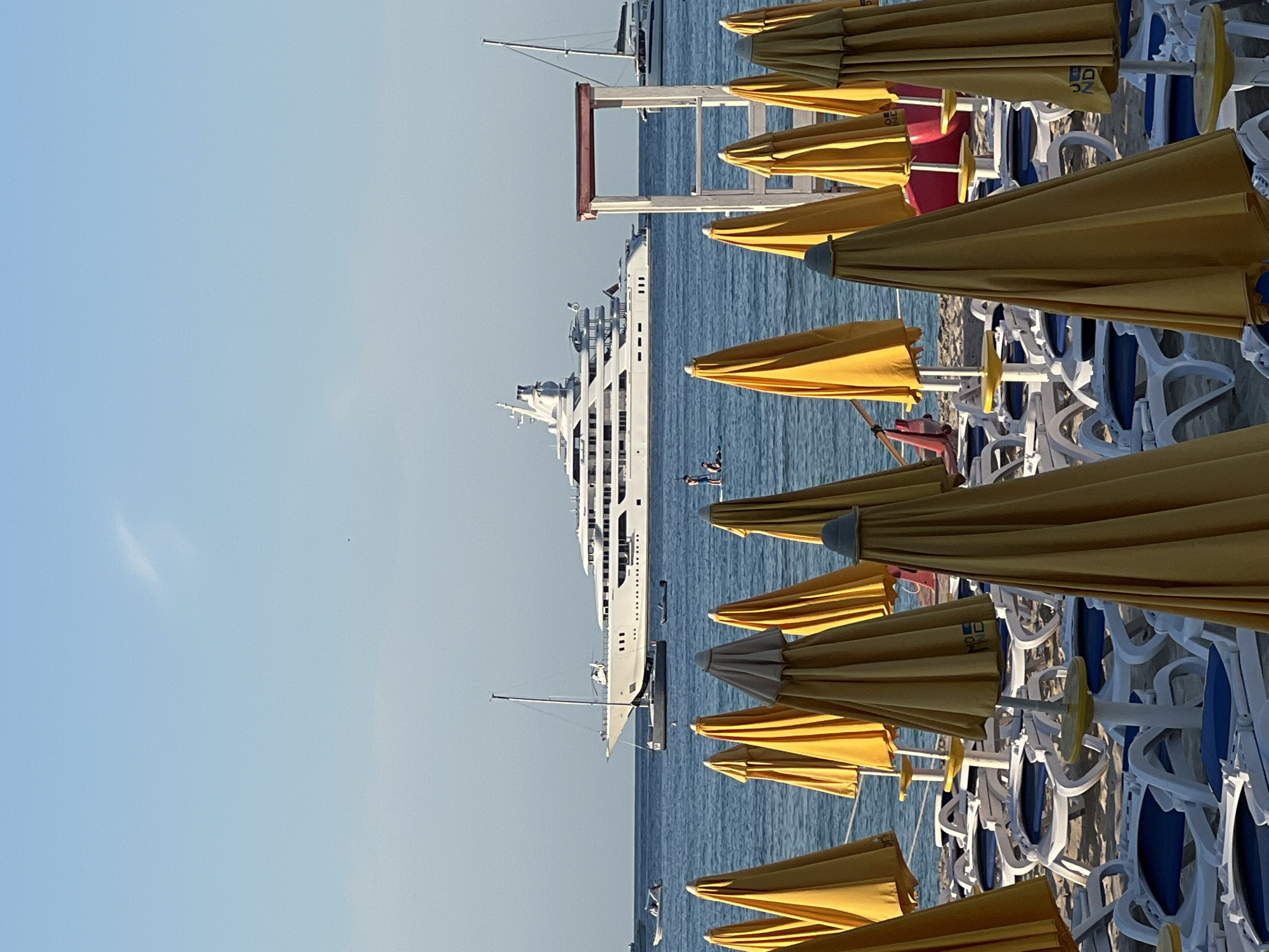 The image depicts a sunny day at a beach, likely in Palermo, Italy, with the focus on a row of closed yellow beach umbrellas and white beach chairs. In the background, a large white yacht is anchored on a calm blue sea. A sailboat is also visible near the yacht, and a smaller boat is further out on the water. The sky is clear and light blue, suggesting fair weather. The umbrellas are densely packed, indicating a popular beach area. Some umbrellas have visible branding, with the text "NOZ" and "DN" discernible on one. The overall scene conveys a peaceful and relaxing atmosphere, with the bright sun casting a warm light on the beach. The time of day appears to be late morning or early afternoon, given the brightness of the sunlight.