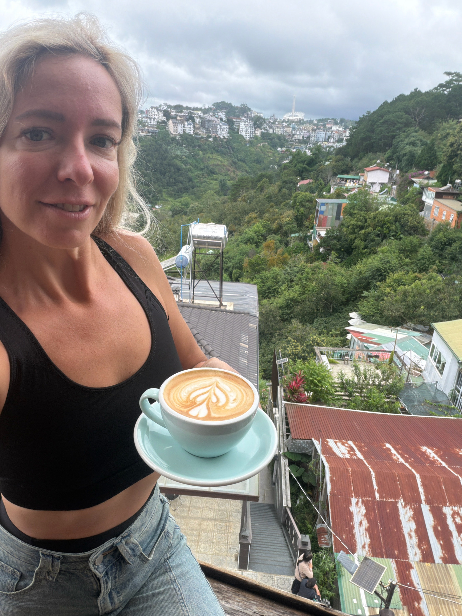 In the foreground, a woman with blonde hair, wearing a black sports bra and light blue jeans, holds a cup of latte with intricate foam art. Her face is partially visible, looking towards the camera with a slight smile. The background reveals a city built on a hillside with lush green vegetation. The city appears to be Da Lat, Vietnam, as indicated by the distinctive architecture of buildings clustered on the slopes and the mountainous terrain. A water tank on a metal frame is visible behind the woman, and a staircase leads down to other parts of the cityscape, where two individuals are seen walking. The sky is overcast with clouds.