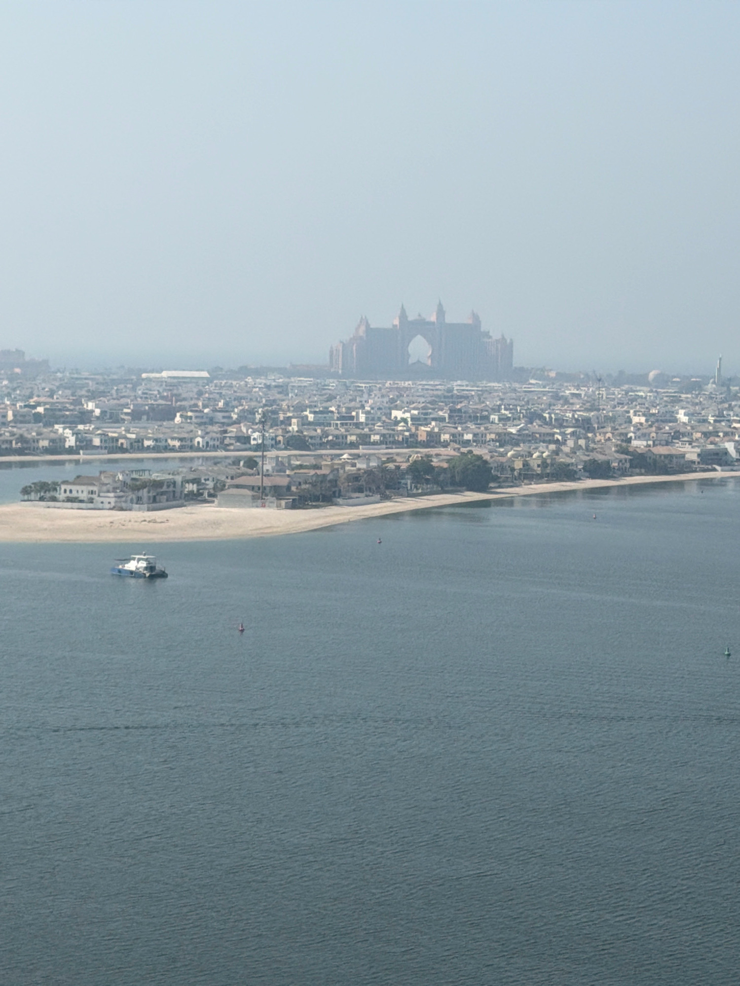The image shows a hazy aerial view of Dubai, United Arab Emirates, featuring a body of water in the foreground and a densely populated coastline with a prominent resort in the background. In the middle ground, a small white and blue boat is visible on the water, positioned near a sandy shoreline dotted with numerous white houses and palm trees. The coastline extends into a city landscape with many buildings of varying heights. In the far distance, the iconic Atlantis, The Palm resort, with its distinctive arch, is partially obscured by a light haze. The water is a calm, blue-gray, and there are several small buoys scattered across its surface. The overall impression is one of a sprawling, sun-drenched urban development adjacent to a tranquil body of water.