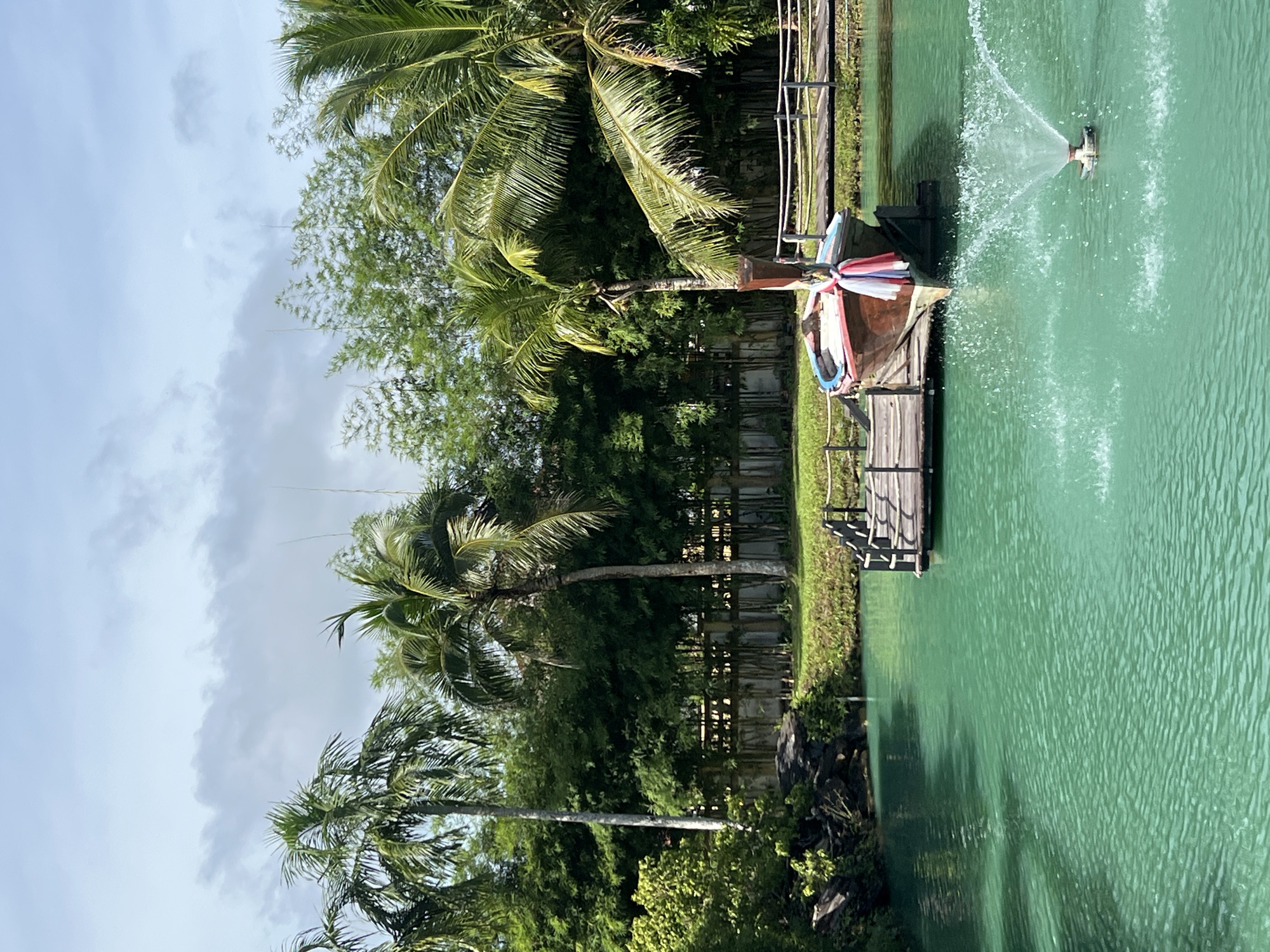 The image depicts a serene waterscape in Tambon Pa Kae Bo Hin, Thailand. The foreground is dominated by a vibrant, turquoise-colored body of water with ripples created by a fountain. Moored to a wooden pier in the middle ground is a traditional Thai longtail boat, adorned with colorful fabric. A wooden bridge stretches across the water to the right of the boat, with a simple wooden railing. The background features lush greenery, including tall palm trees and bamboo stalks, against a cloudy sky. The scene is tranquil and idyllic, suggesting a peaceful natural setting within the specified location.