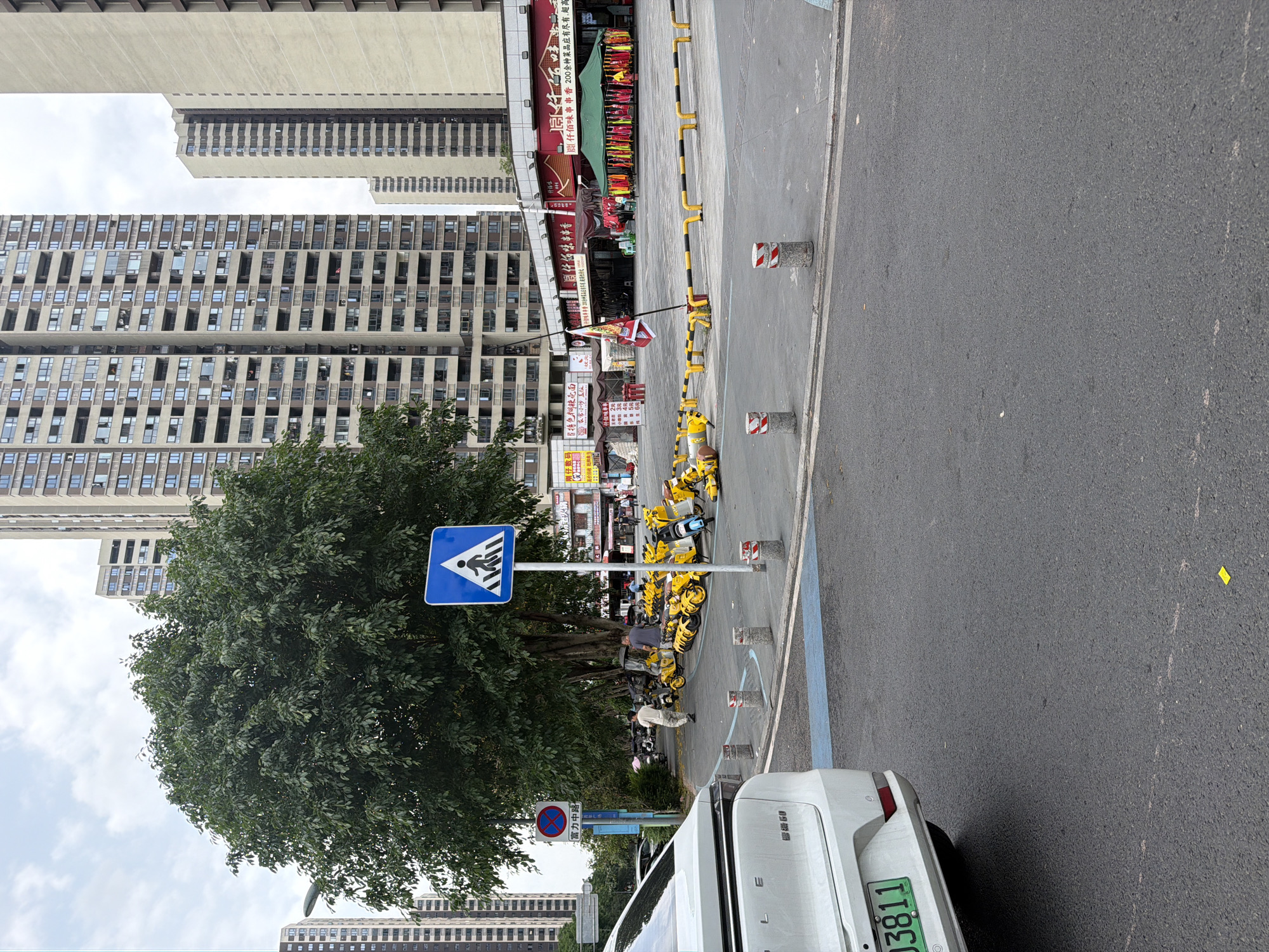 The image depicts a street scene in Chongqing High-tech Zone, China. In the background, tall residential buildings are visible, alongside commercial establishments, one with a red awning and Chinese characters. A pedestrian crossing sign is positioned near a large tree. Several yellow shared bicycles are parked nearby. A white car is partially visible in the foreground, with the license plate “E03811”. Street infrastructure, including poles and concrete bollards, is present. Two people are walking near the parked bicycles. The environment appears to be an urban area with commercial and residential activity.