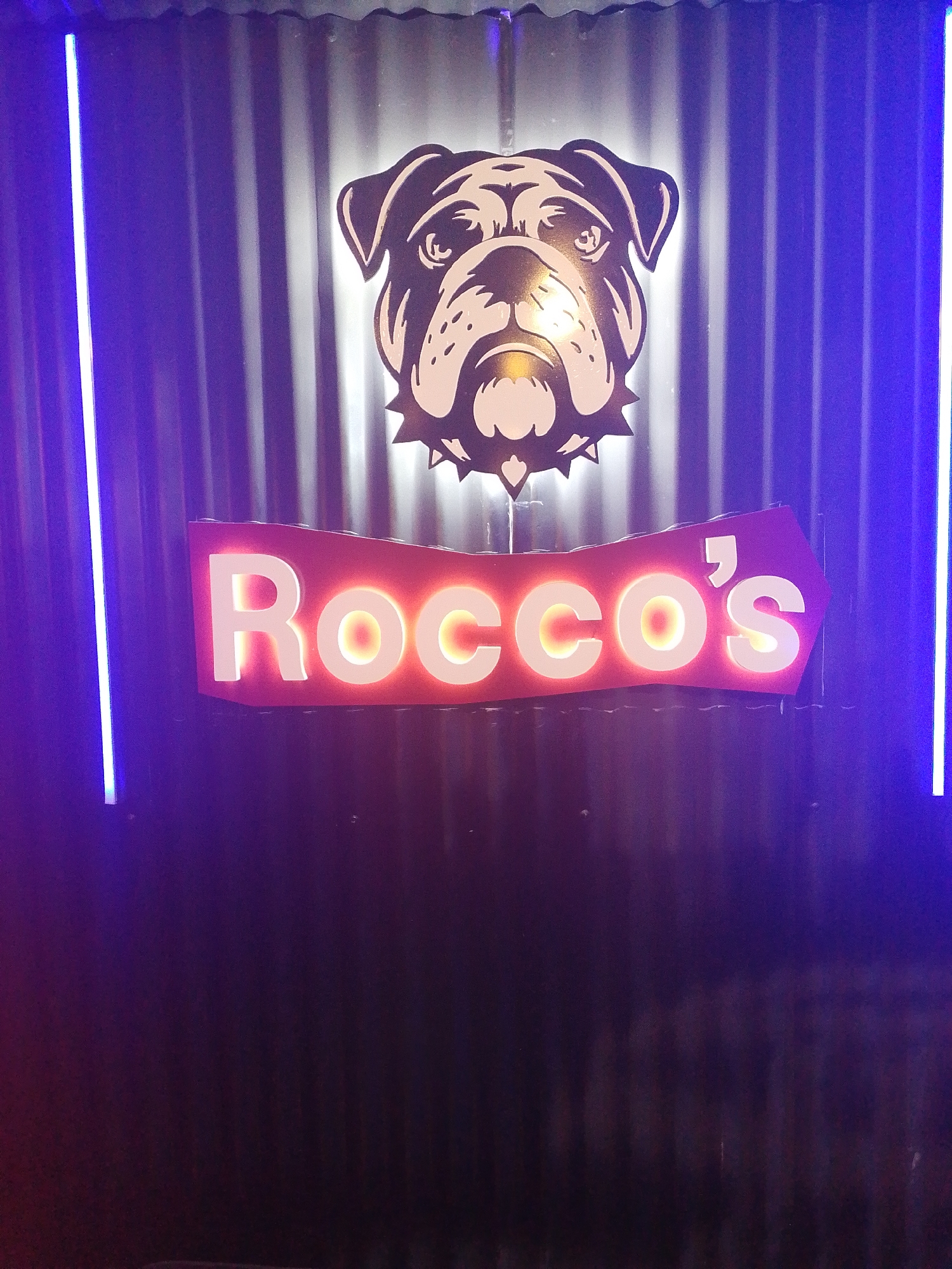 The image shows a sign for "Rocco's" with a bulldog head logo above it. The sign is illuminated, with the letters appearing to glow. The background is a corrugated metal wall. Two vertical blue lights flank the sign. The location is presumably Tinaquillo, Venezuela. No people or interactions are visible in the image.