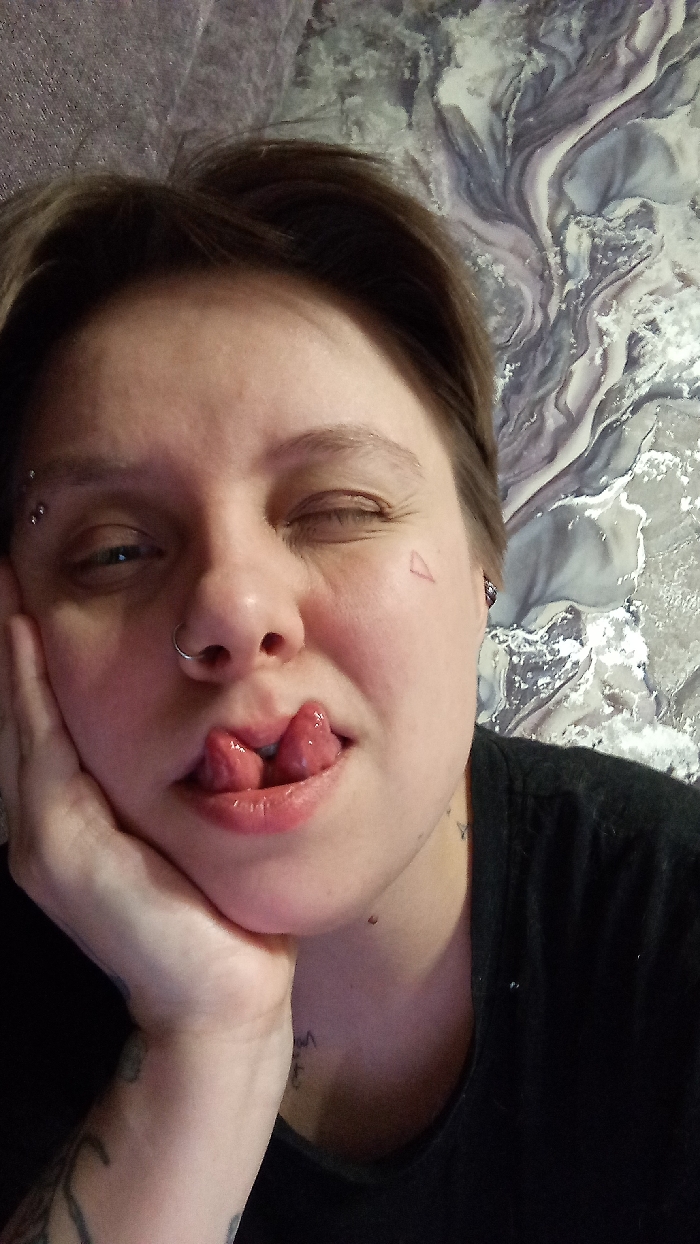 This is a close-up, front-facing portrait of an individual, likely captured indoors in Saratov, Russia. The subject is an adult with fair skin and short, dark brown hair. They are winking with their right eye (viewer's left) while their left eye is partially open and looking slightly to the right. Their bifurcated (split) tongue protrudes from their slightly open, glossy lips. The individual's left hand is positioned to support their chin and jaw.
Visible body modifications include two barbell piercings on the left eyebrow, a silver hoop nose ring in the left nostril, and a small stud or ring in the right earlobe. A small red outline of a triangle is tattooed on the right cheekbone. Faint script tattoos are visible on the neck/collarbone area, and a patterned tattoo is partially visible on the left forearm/upper arm.
The subject is wearing a plain black t-shirt. The background consists of a wall featuring a marbled or swirling pattern in shades of grey, white, and light purple/blue, with a section of what appears to be a textured, possibly padded, dark grey surface visible in the upper left quadrant. The lighting appears to be ambient indoor light. The specific location within the city of Saratov cannot be determined from the image.