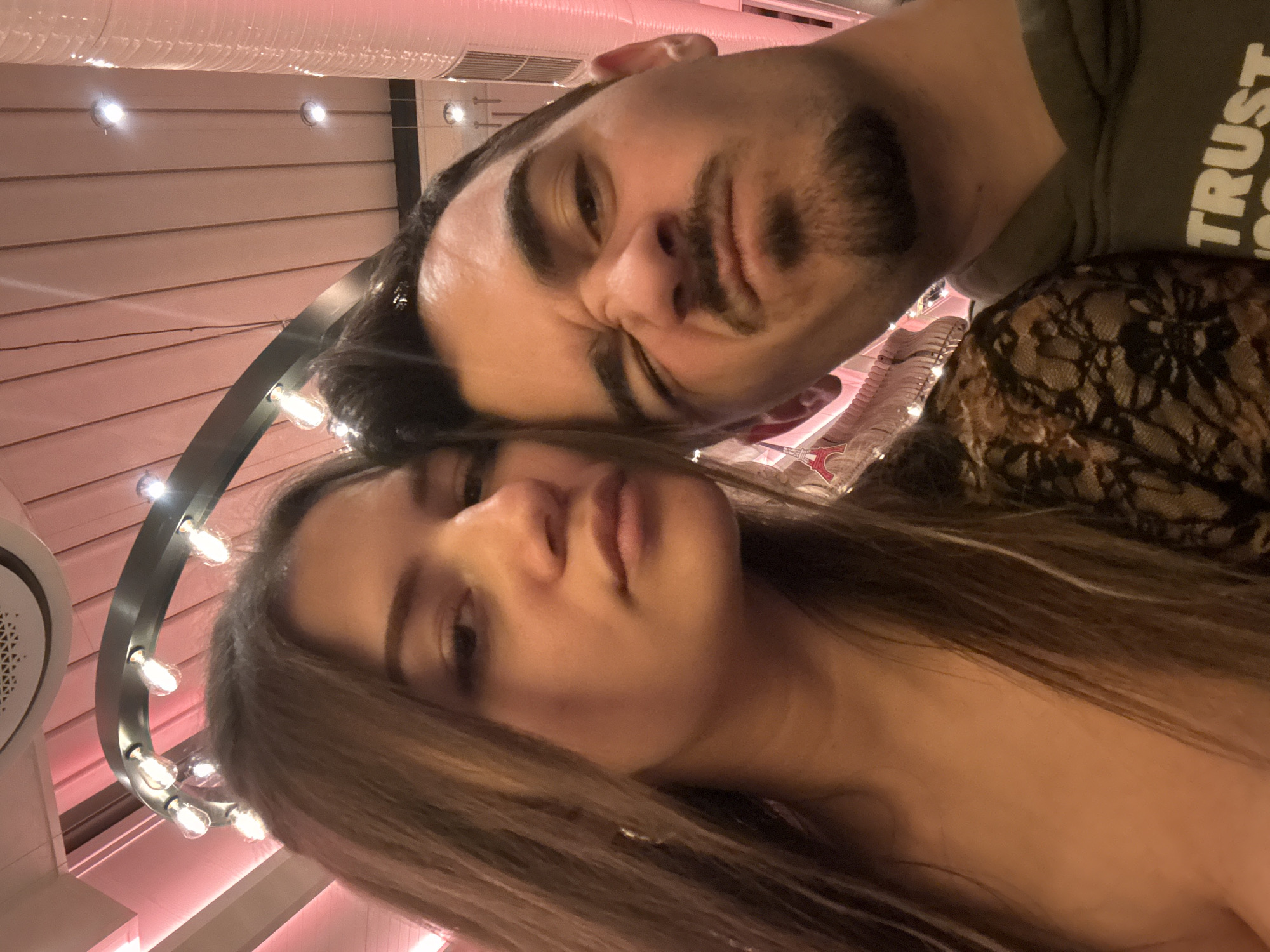 This image captures a close-up selfie of two individuals, an adult female and an adult male, within an indoor commercial establishment in Sofia, Bulgaria. The environment strongly suggests a modern restaurant or cafe, consistent with a "Happy Bar & Grill" location, characterized by specific interior design elements.
The female, positioned on the left, has long, straight, dark brown hair and displays a neutral, direct gaze at the camera. She is wearing a black lace garment, with a partial gold-colored hoop earring visible on her left ear.
The male, positioned on the right and slightly angled closer to the camera, has short dark hair, a mustache, and a goatee. He exhibits a playful expression, with his right eye tightly squinted and his lips pursed. He wears an olive-green hooded sweatshirt, with white text, partially reading "TRUST", visible on the lower right chest area.
The background features a distinctive ceiling structure with light pink/red corrugated or fabric-lined panels and white sections. Prominent lighting fixtures include an arched dark-colored metal bar with multiple illuminated incandescent-style bulbs, and several small, bright recessed spotlights. A translucent red, tube-like structure, possibly a decorative air duct, is visible vertically on the far right. A small, red Eiffel Tower silhouette decoration is present in the background below the male's right ear. The overall lighting is dim and atmospheric, characterized by warm pink and red hues.