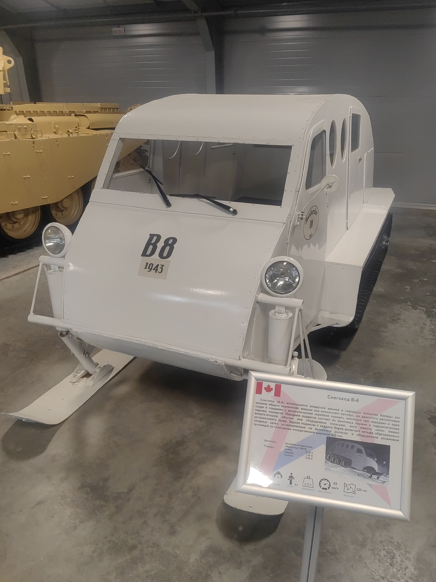 This media file is a photograph of a white, retro-style snowmobile, identified by the text on its side as "B8 1943." The snowmobile is positioned indoors, likely in a museum or exhibition space, as indicated by the smooth concrete floor and the gray, corrugated wall in the background. To the left of the snowmobile, a portion of a larger, yellow military vehicle, possibly a tank, is visible.
The snowmobile has a distinct, rounded cabin with several circular windows. It features two prominent headlights at the front, a windshield with wipers, and skis at the front for locomotion. A continuous track system is visible at the rear.
In the foreground, a silver-framed information sign is placed on a stand. The sign displays a Canadian flag and text in Russian, identifying the vehicle as "Снегоход В-8" (Snowmobile B-8). The text provides details about its use by the Canadian army in northern regions, its engine, dimensions, and capabilities, including speed and range.
The overall impression is one of a well-preserved historical artifact displayed in a controlled environment, likely part of a military or transportation museum. The lighting is artificial and even, suggesting an indoor setting. No people are visible in the image. The setting appears to be a museum in Kubinka, Russia, based on the provided location context.