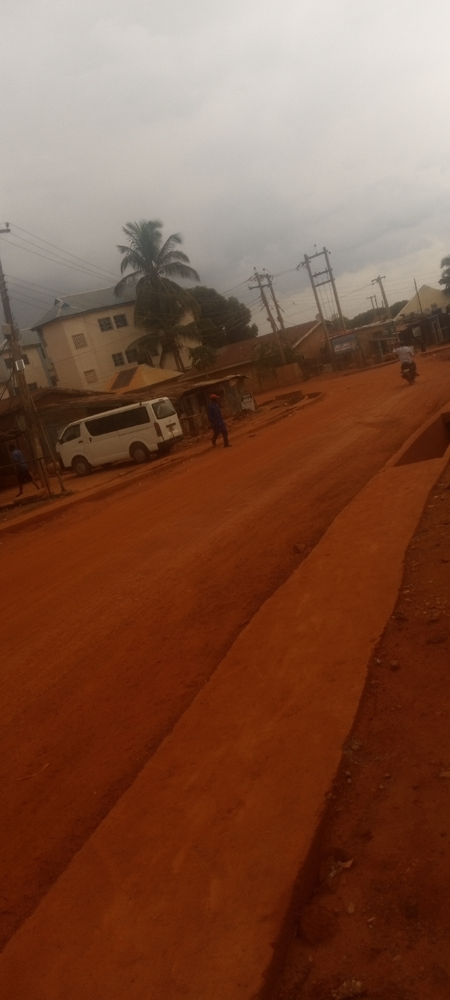 The image depicts a street scene in Yelwa, Nigeria, under a cloudy sky. The ground is red dirt. On the left, a white van is parked near a two-story white building. Power lines and poles run overhead. A person in blue clothing walks on the left side of the street, while another person rides a motorcycle to the right. A concrete curb runs along the right foreground.
