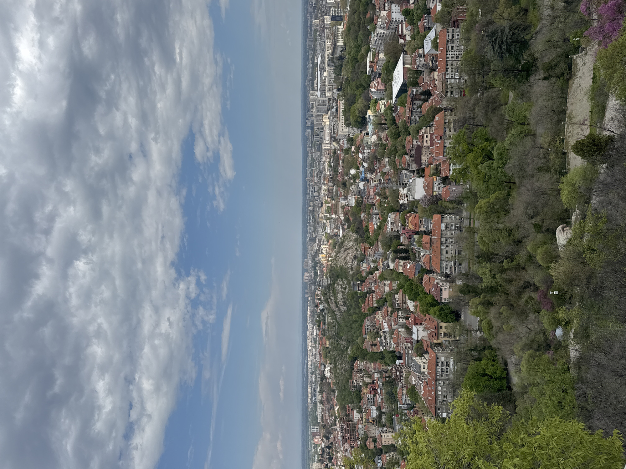 This wide-angle photograph presents a panoramic view of the city of Plovdiv, Bulgaria, captured from a high vantage point, likely one of its iconic hills. The image is horizontally divided, with the upper half showcasing a partly cloudy sky and the lower half revealing the extensive urban landscape.
The sky displays a mix of bright blue patches and scattered, fluffy white and grey clouds, suggesting fair weather during the daytime, possibly late morning or early afternoon. Below the horizon line, the city sprawls outwards, densely packed with buildings that stretch into the distance. Most structures are multi-story, characterized by their reddish-orange tiled roofs, a common architectural feature in the region.
Throughout the cityscape, vibrant green trees and wooded areas are interspersed among the buildings, particularly noticeable on the slopes descending from the viewpoint. Some trees exhibit fresh, bright green foliage of spring, while others appear more dormant or show hints of purplish blossoms. In the mid-ground, a distinct, large white building with a flat, somewhat triangular roof stands out against the residential backdrop. Closer to the foreground on the right, rough rock formations and a section of a light-colored stone or concrete wall/path are visible, likely part of the hill's natural terrain or infrastructure. The immediate foreground on the left is framed by soft, green leaves, indicating the shot was taken from within or behind some foliage. There are no visible people or discernible text in the photograph.