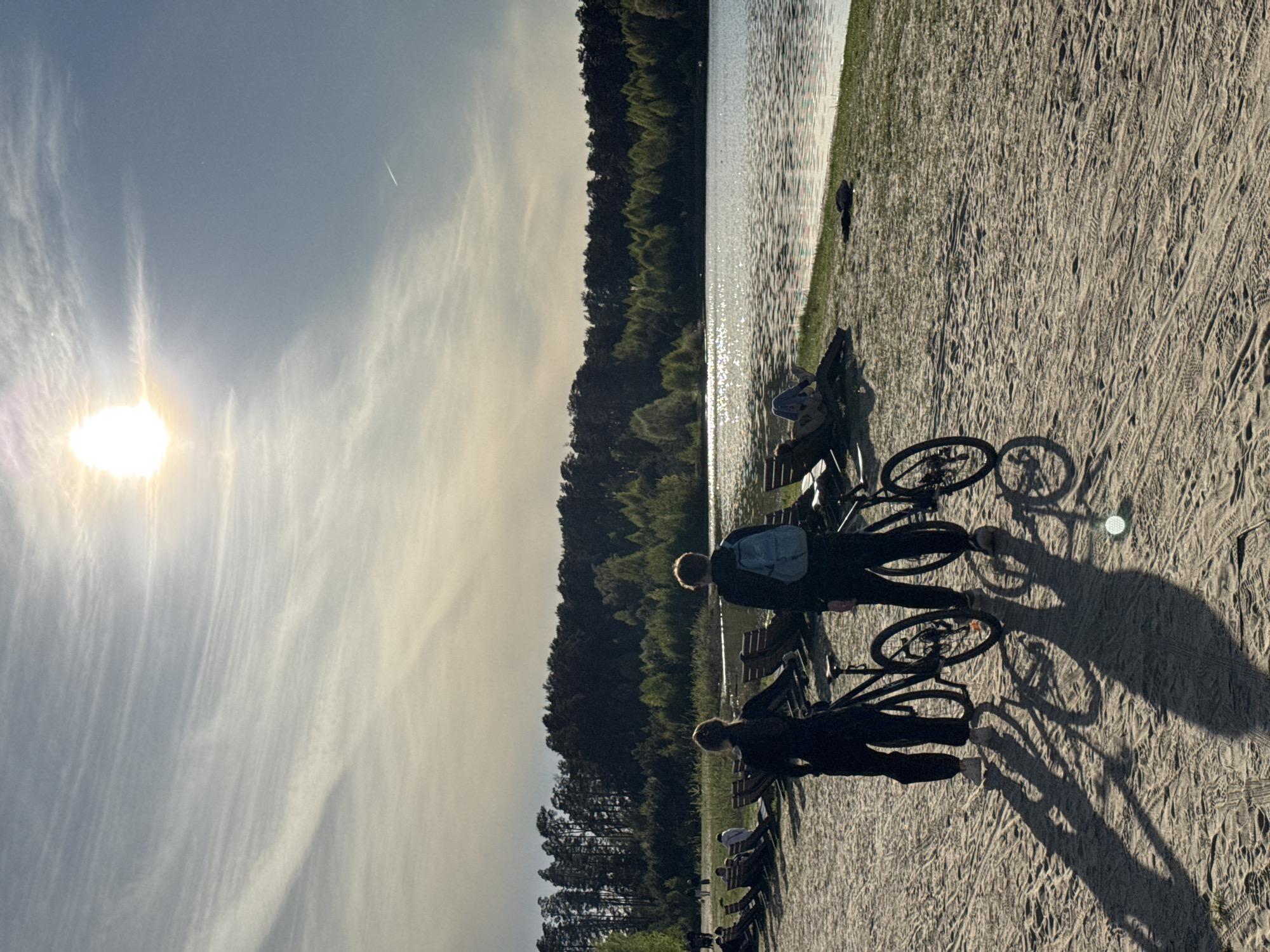 The image shows a lakeside scene in Kazan, Russia, during daylight. Two people, possibly a man and a woman, stand with their bicycles on a sandy area next to a body of water lined with trees. The sun is visible in the sky, creating shadows of the people and bicycles on the sand. There are benches along the sandy area. The lake's surface reflects light.
