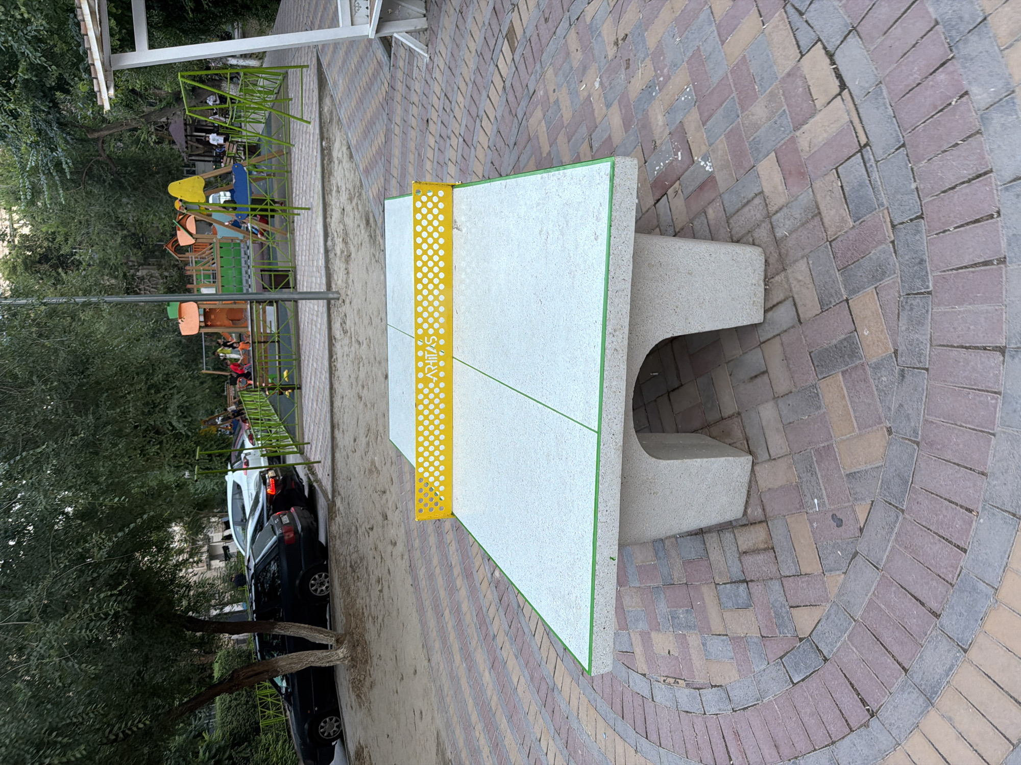 In Shymkent, Kazakhstan, a concrete ping pong table with a yellow net stands on a patterned brick patio. In the background, a playground with swings and slides is visible, occupied by several children and adults. Cars are parked to the left of the playground, near trees and a fence. The overall environment appears to be an outdoor recreational area within a city.