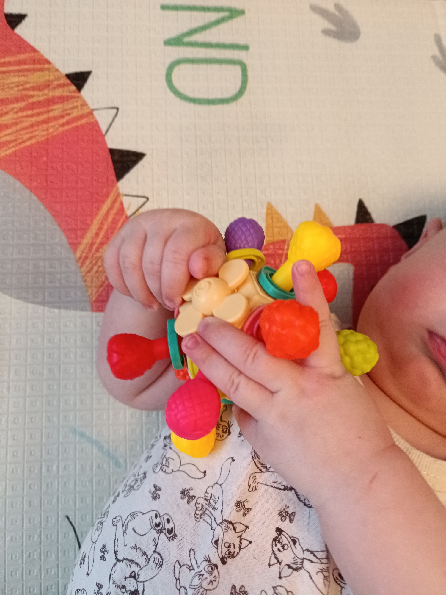 A baby lying on a play mat is holding a colorful baby toy. The mat has a dinosaur design and the word "NO". The baby is wearing white pajamas with a black cartoon animal print. The location is not discernible from the image.