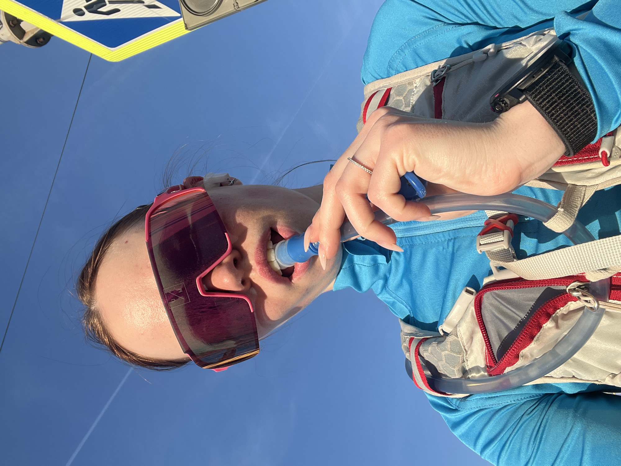 A woman in Chaikovsky, Russia, wears pink sunglasses, a blue shirt, and a hydration pack. She drinks from the hydration pack's tube. A sign with a pedestrian crossing symbol is visible in the background. The sky is blue and clear.