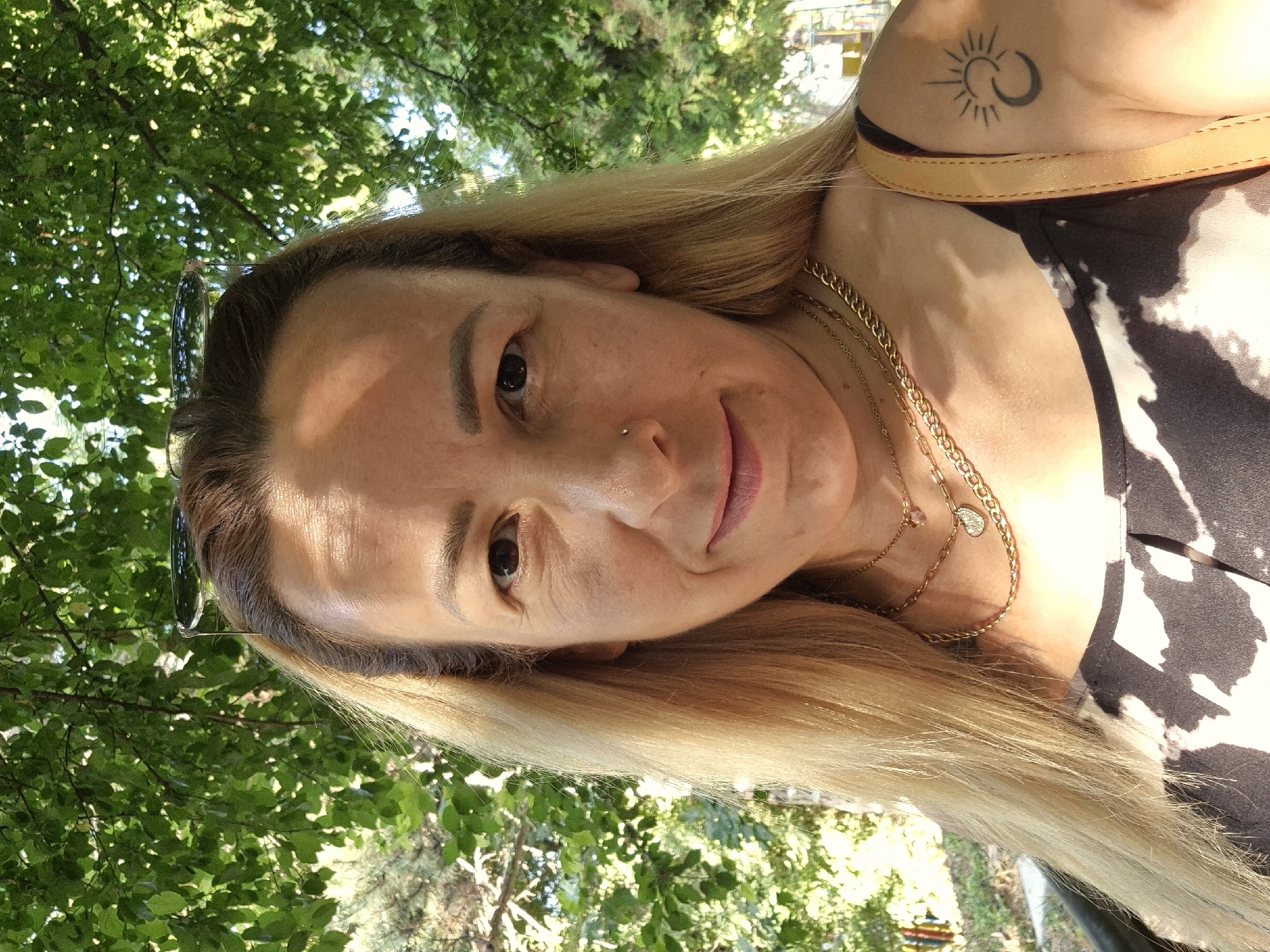 A medium shot, outdoor, selfie shows a woman with blonde hair and a nose ring looking at the camera. She is wearing two gold necklaces, one with a small pendant and another with a heart-shaped pendant. She has a tattoo of a sun and moon on her left shoulder. She is wearing a black and white patterned top. Behind her is a lush green tree canopy with dappled sunlight filtering through. The background is blurred, but hints of trees and possibly a fence can be seen. Sunglasses are perched on her head.
