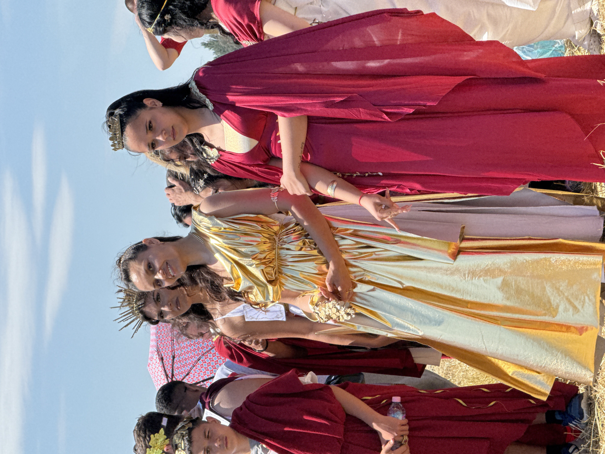 The image depicts an outdoor scene in Gigen, Bulgaria, under a clear blue sky with bright sunlight. The ground is covered with dry, yellowish-brown grass.
The foreground is dominated by two women dressed in elaborate historical costumes. The woman on the left wears a shimmering golden, full-length gown with flowing drapes. She has dark hair, a subtle necklace, and a small, decorative, gold-colored cluster of objects in her left hand. She is smiling towards the viewer, and her right hand holds the left arm of the woman next to her.
The woman on the right wears a deep red, full-length gown with gold trim along the neckline and an attached cape-like drape. She has dark hair, pulled back, and wears a gold crown/headband. Her expression is pensive, and her left arm is held by the woman in gold. A faint tattoo is visible on her right forearm.
To the far left, a young boy, appearing to be pre-teen, is partially visible. He wears a red toga-like garment and a gold leaf-patterned headpiece. He holds a pink plastic water bottle in his left hand.
Behind the central figures, several other individuals are partially visible, some also in historical attire. A woman with dark hair and a distinct spiky gold crown is seen behind the woman in gold. Further back, a red umbrella with a white geometric pattern provides shade. On the far right, another woman in a red garment with a gold headpiece is partially visible.