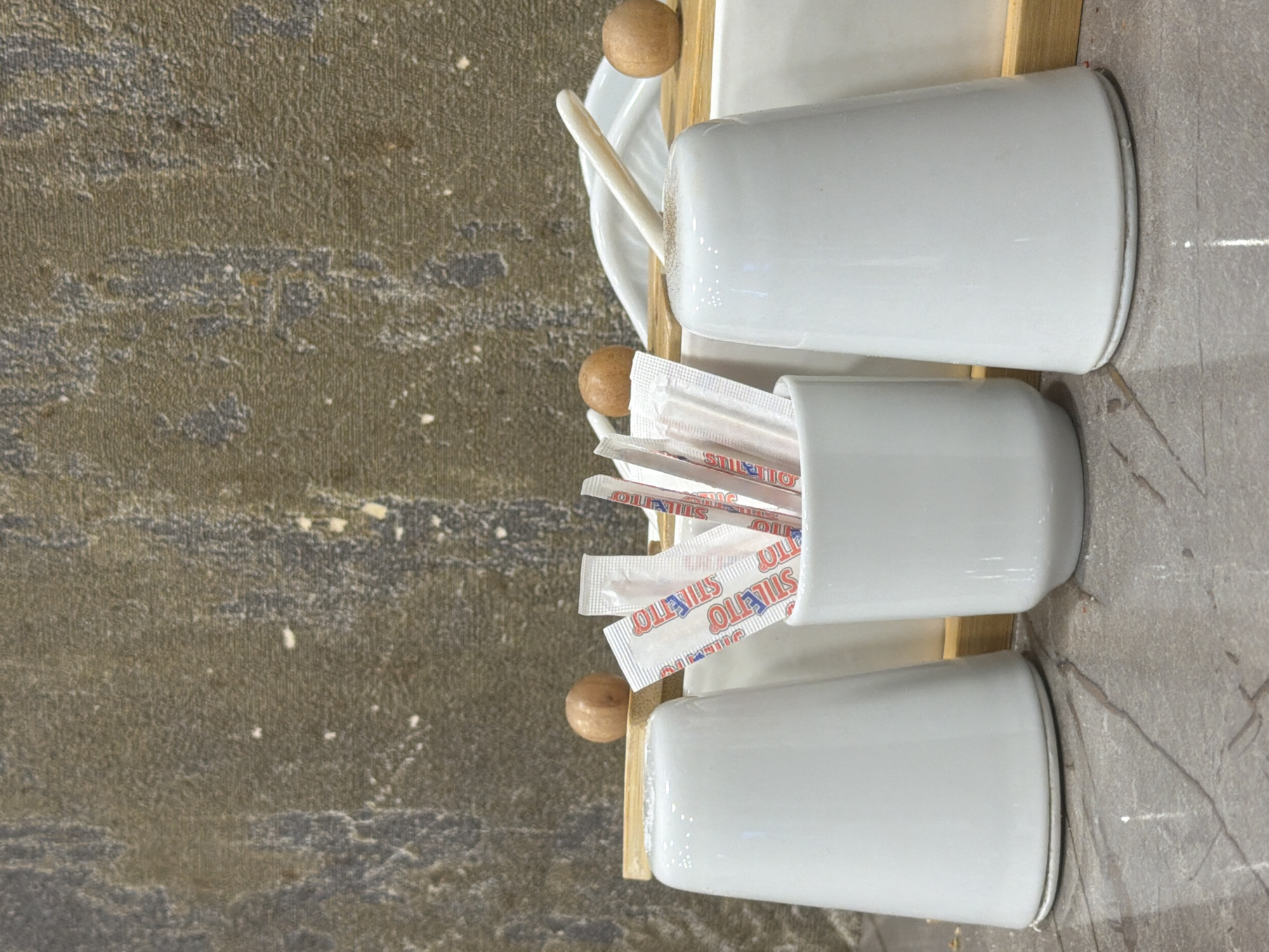 A top-down, close-up shot reveals a wooden tray holding three white ceramic containers and a spoon, sitting on a surface composed of two different textures. To the left is a mottled, somewhat rough, grey-brown surface, while to the right is a smoother, grey-white surface with faint, dark lines resembling veins. The containers are arranged linearly. The first container, positioned at the top of the frame, holds a long white spoon and is sitting on a white saucer. The second container is filled with packets of white paper straws with red printed lettering. The third container is empty. All three ceramic containers are white with a cylindrical shape. The wooden tray is a light color, and round wooden knobs are attached to the corners of the tray. The scene is captured with static camera work and neutral lighting. There are no people in the frame. The location is inferred to be within Podgorica, Montenegro, likely inside a cafe or restaurant setting based on the objects present, but precise geolocation is not possible from the visual data alone.