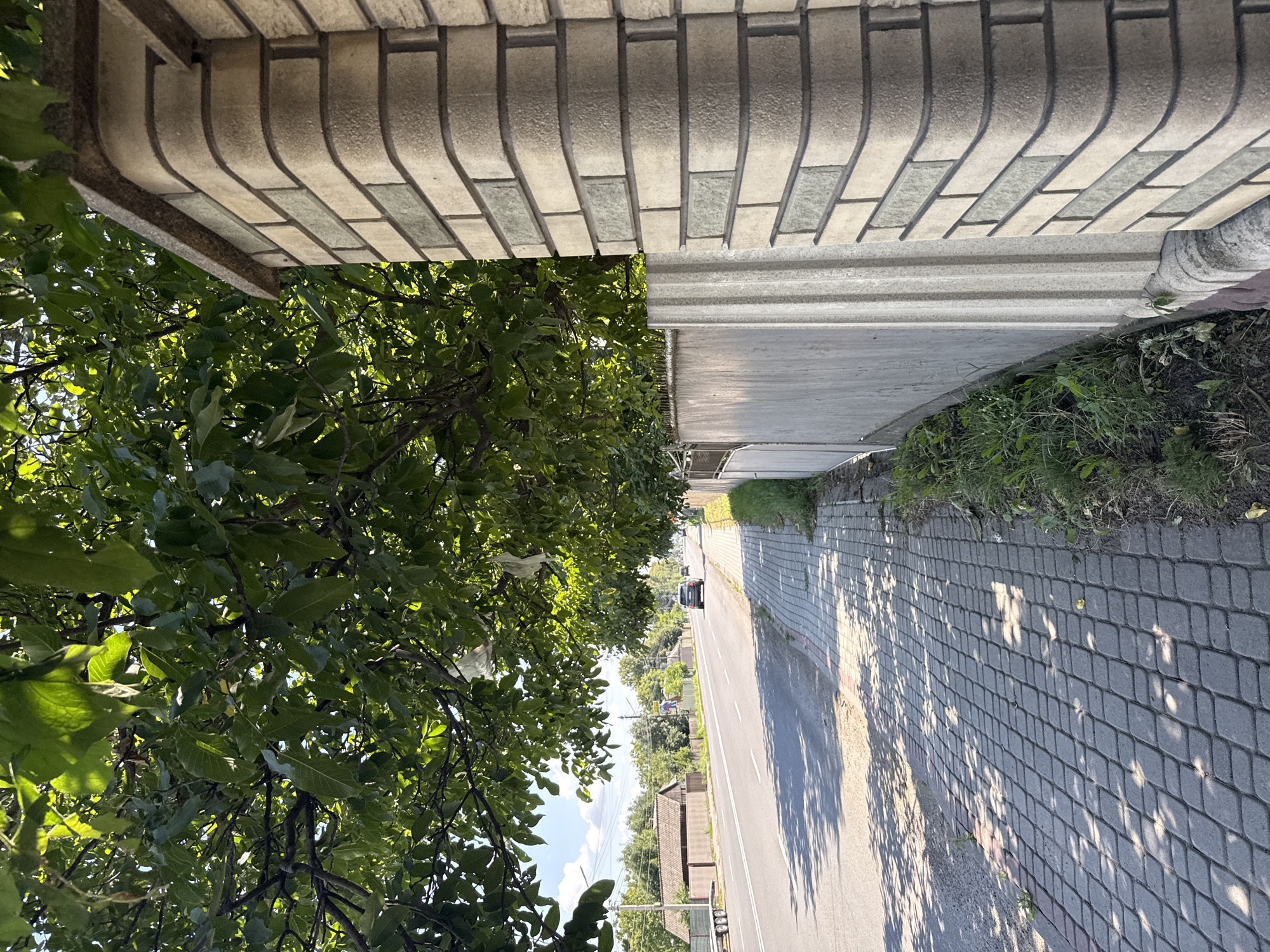 An eye-level, outdoor, medium shot shows a paved sidewalk and a road on a sunny day. In the foreground, a brick pillar with a curved edge and horizontal bands of light and dark grey bricks stands on the right. To the left of the pillar is a light grey concrete fence with vertical grooves. A tree with green leaves hangs over the sidewalk and road, casting dappled shadows on the paving stones. The road, a two-lane asphalt surface with white dividing lines, extends into the distance. A dark-colored car is visible driving on the road. On the left side of the road, there are houses with brown roofs, a tall green tree, and a blue sky with white clouds. The sidewalk is made of grey rectangular pavers laid in a diamond pattern. Green grass and weeds grow along the base of the concrete fence.