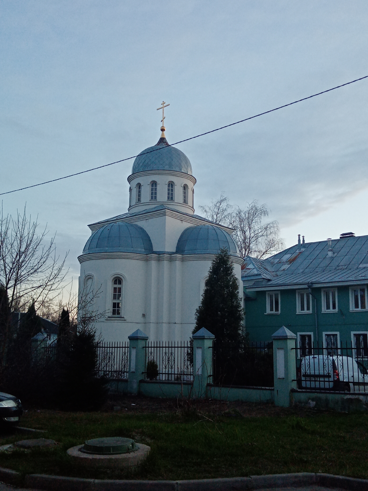 The image depicts a white Orthodox church with blue domes and a gold cross in Elektrostal, Russia. The church is surrounded by a dark metal fence with green posts. In the background, there is a green two-story building with a gray roof. The foreground includes a patch of grass and a concrete curb. A black car is partially visible to the left of the church. A utility wire crosses the sky diagonally. The sky is overcast.