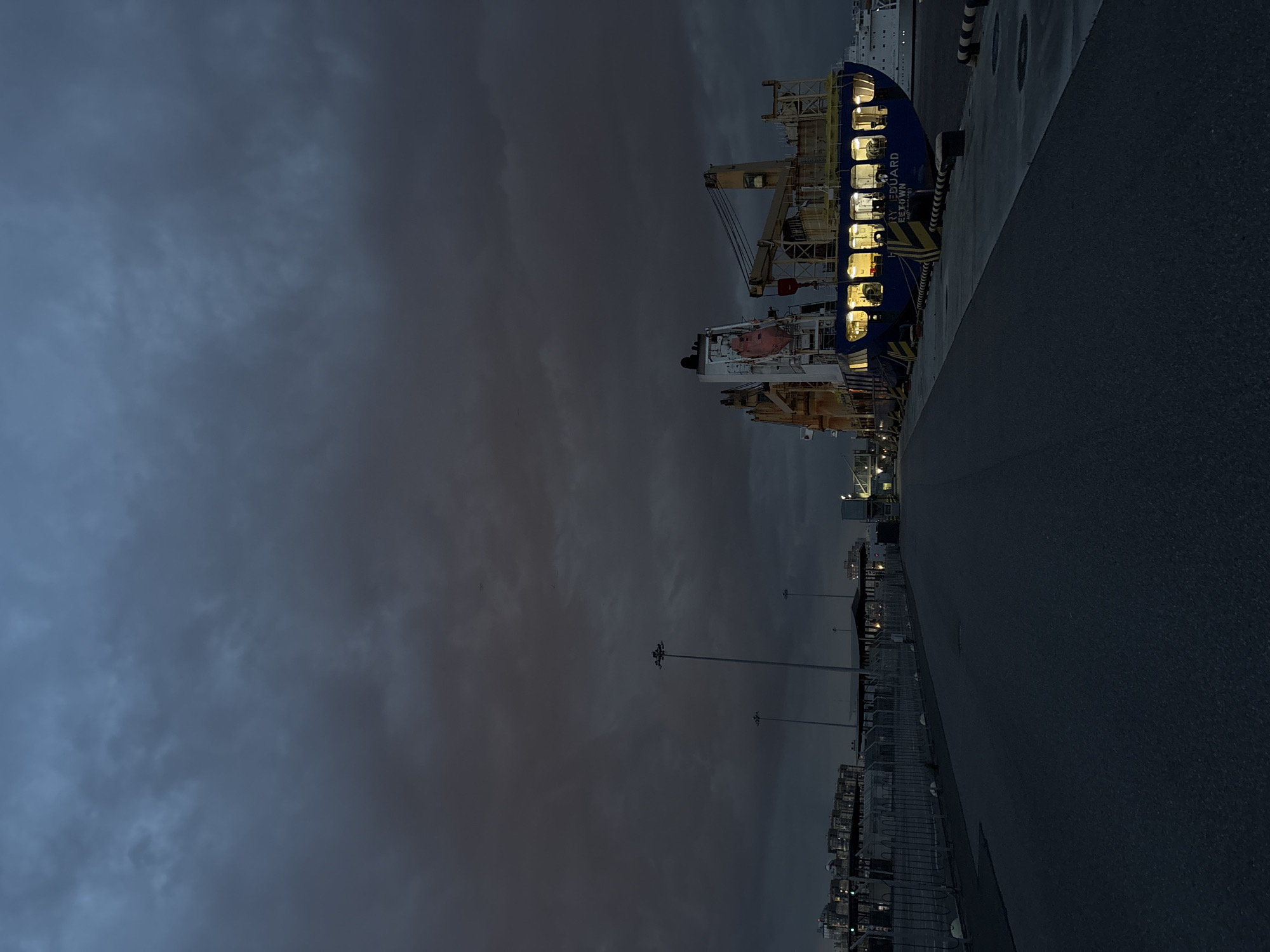 A cargo ship named "HERRY EDUARD" is docked at a pier in Saint Petersburg, Russia. The ship is illuminated from within, with windows showing warm light. The sky is dark and cloudy, suggesting it is either dawn or dusk. A paved road leads to the pier, with a fence and lampposts lining the side. Other buildings and structures are visible in the background. The scene appears to be a port or industrial area.