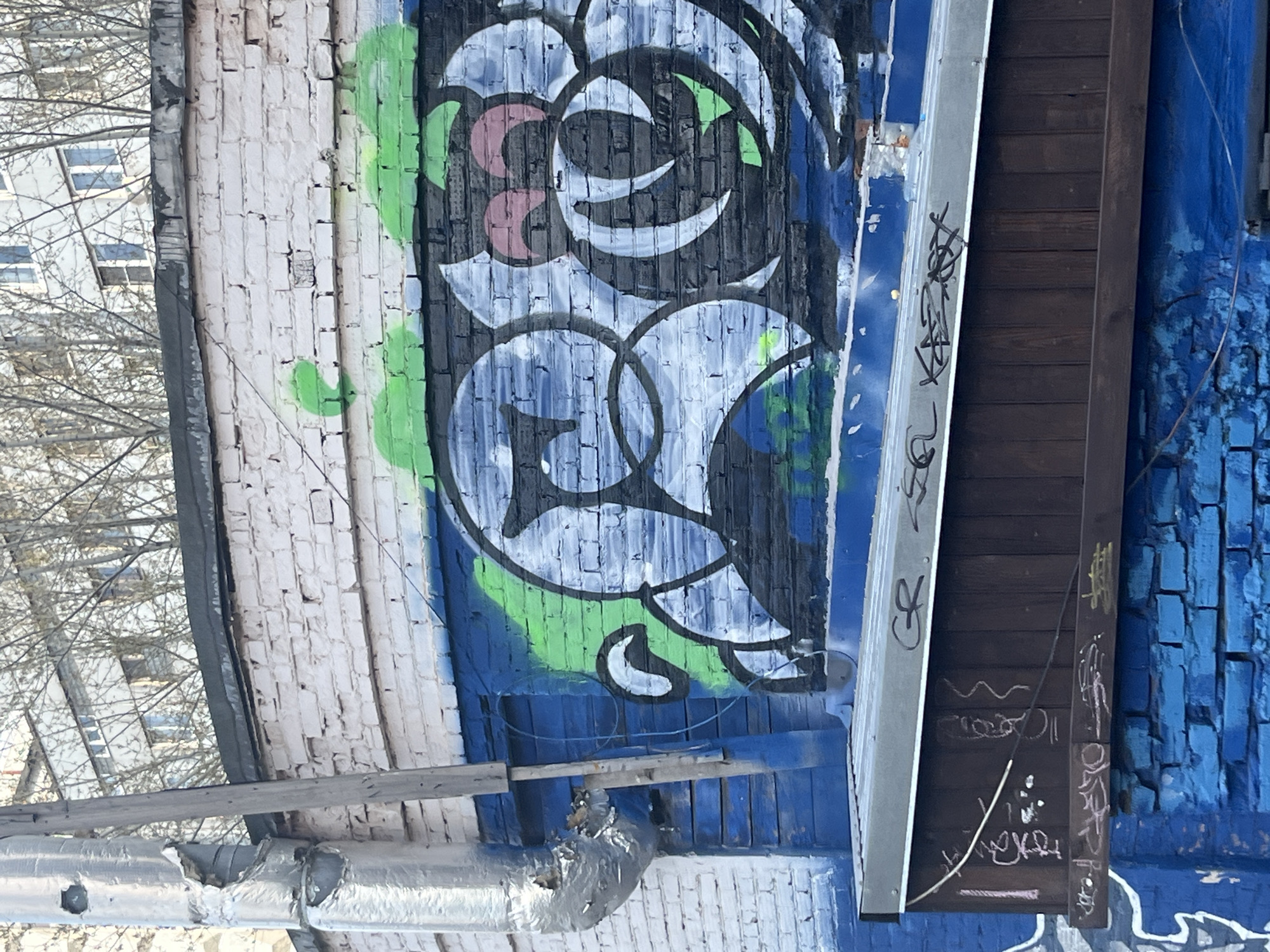 The image shows a section of a brick wall covered in graffiti, likely located in Krasnoyarsk, Russia. The wall is painted in various colors, including white, blue, green, and black. The graffiti consists of abstract designs and tags. A wooden structure with more graffiti on it is attached to the wall. A silver pipe runs along the bottom of the wall. In the background, a building is visible through bare tree branches.