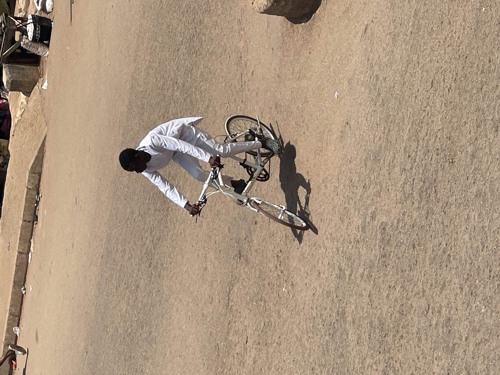 A person dressed in white is riding a white bicycle with red rims on a dirt road. The person is looking to the left. The bicycle has a black seat and black handlebars. The person is wearing grey shoes. The road is dry and dusty. In the background, there are some structures that appear to be market stalls, with bags and various objects on display. There is also a curb with some litter on it. The scene is lit by sunlight, casting shadows on the road.