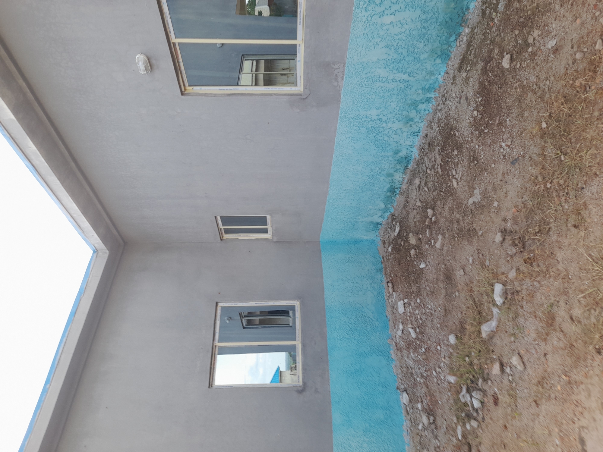 The image shows the exterior of a building under construction, likely in Ibi, Nigeria. The building has grey plastered walls and a blue textured base. There are two windows visible on the left side of the building and a larger window on the right, all with white frames. The ground in front of the building is covered with loose soil, rocks, and some sparse vegetation. The sky is visible at the top of the image. The area appears to be an open space adjacent to the building, possibly a construction site or an undeveloped plot of land. There are no people or animals visible in the scene.