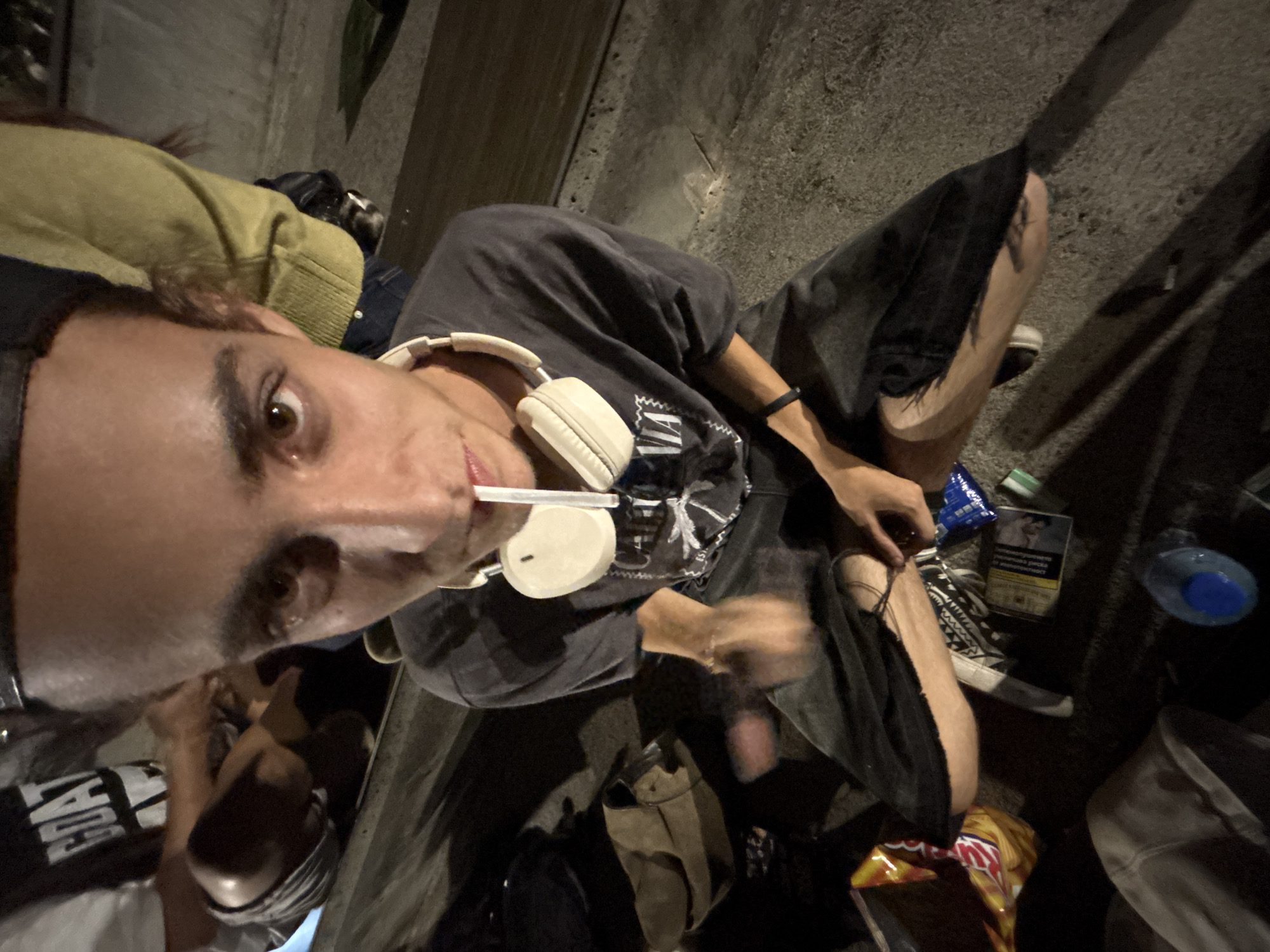 A person is sitting on a concrete bench outdoors at night. They are wearing a dark t-shirt, dark pants, and a black baseball cap. A pair of white headphones are around their neck. A cigarette is held between their lips. To their left, a bag is on the ground, and to their right, there are several items including a pack of cigarettes, a blue bottle cap, and other assorted items. The background is dark, suggesting it is nighttime. The location is identified as the city of Vratsa, Bulgaria.