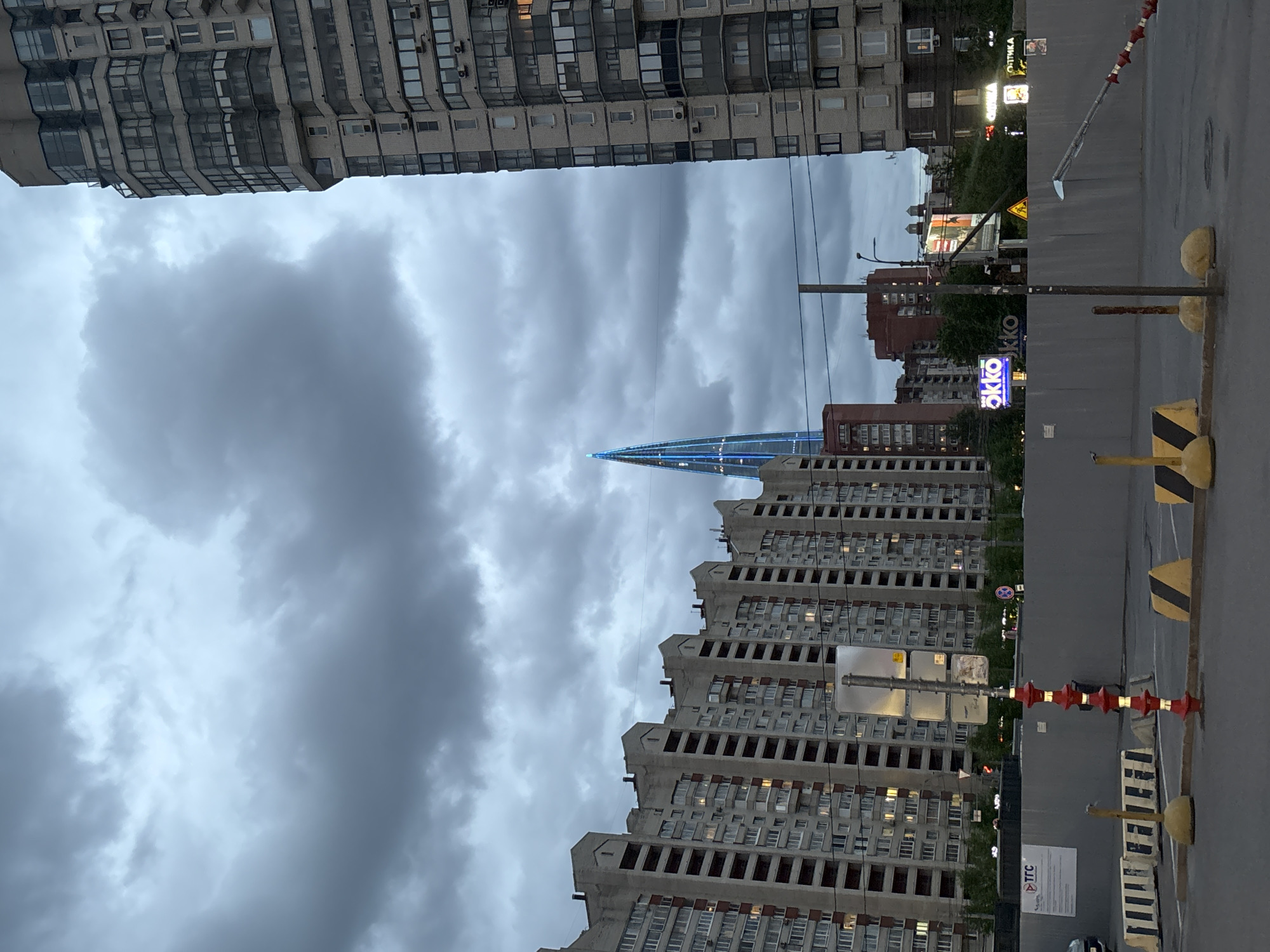 The image shows a view of Saint Petersburg, Russia, on a cloudy day. Two large apartment buildings frame the sky. A taller, modern skyscraper with a blue-lit spire is visible between the buildings. A street runs in the foreground with construction barriers on the right. A sign with "OKKO" in blue neon is visible. Other signs in Russian are also present.