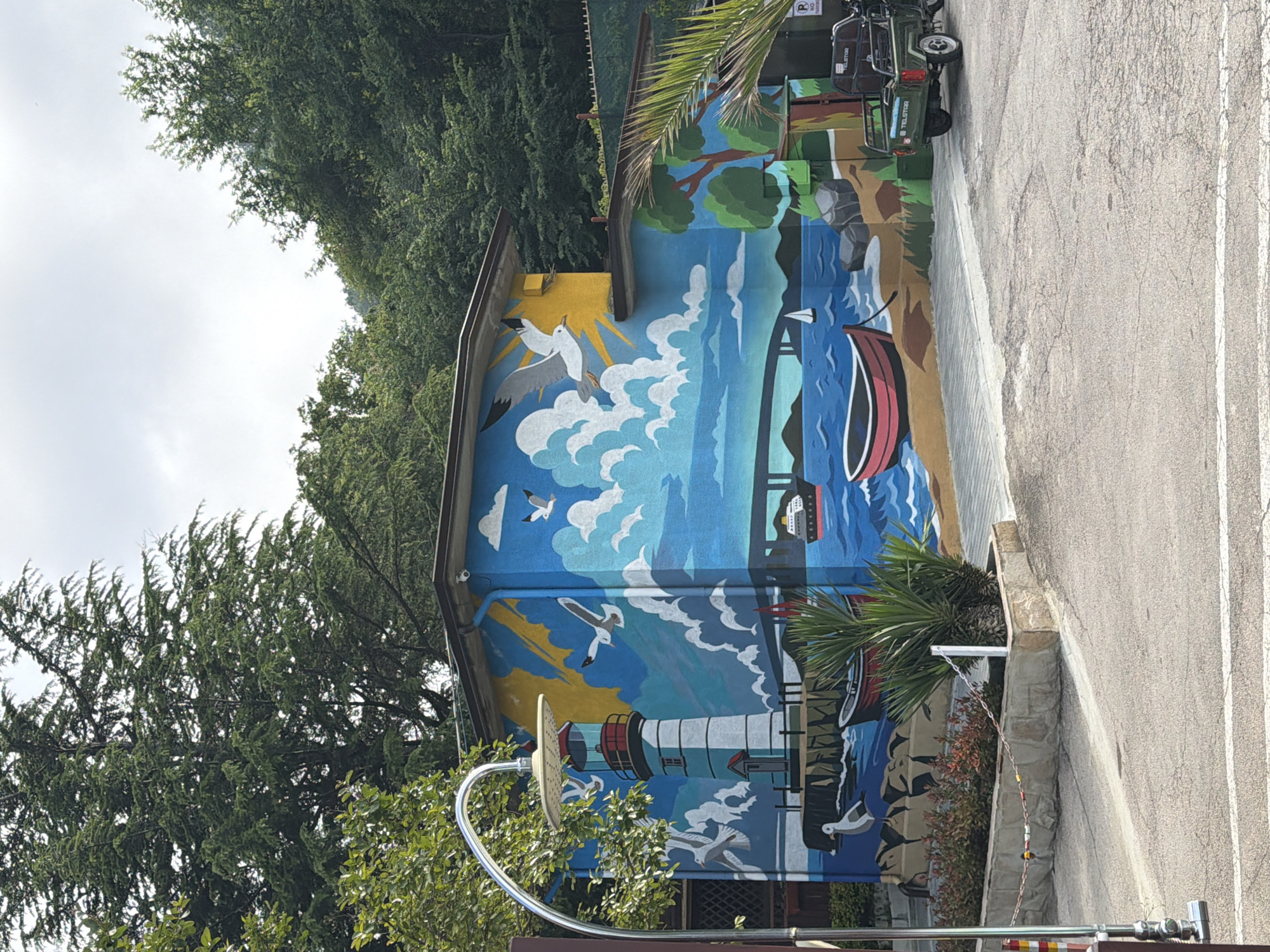 This media file captures a building adorned with a vibrant mural, set against a backdrop of lush green trees and an overcast sky, likely in Varna, Bulgaria. The image is oriented vertically (rotated 90 degrees clockwise).
The central subject is a two-story building with a large, colorful mural painted across its side. The mural depicts a scenic coastal landscape, featuring a blue sky with white clouds and a bright yellow sun. Numerous white seagulls are painted in flight against the sky and above the water. A prominent white and red lighthouse stands on a rocky shore, casting its light. Below the lighthouse, a blue body of water, possibly the sea or a lake, has white waves and several boats, including a red and white rowboat and a white sailboat. The shoreline is depicted with brown earth and green foliage, including palm trees.
To the left of the building, a dense canopy of green trees fills the upper portion of the frame. In the foreground, near the bottom of the building, a metallic outdoor shower fixture with a round head is visible, partially obscured by green bushes. To the right of the building, a paved asphalt area, likely a parking lot or road, is visible, with a white painted line. A small, dark green utility vehicle or golf cart, with "TELSTAR" written on its side, is parked near the building. A partially visible parking sign with a white "P" on a blue background is also seen. The time of day appears to be daytime, with soft, diffused light suggesting an overcast sky. No people are visible in the image.