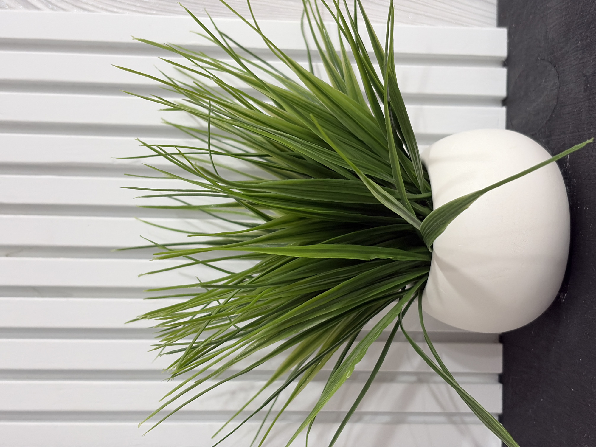 A close-up shot shows a potted faux grass plant on a dark surface in front of a white, slatted background. The plant has numerous long, thin, green blades that fan outwards from a white, rounded pot. The background consists of vertical white slats that create a ribbed texture. The lighting is even, highlighting the texture of the slats and the vibrant green of the plant. The scene appears to be indoors, possibly in a home or office setting, with no discernible urban or natural elements. The specific location within a city is unknown.