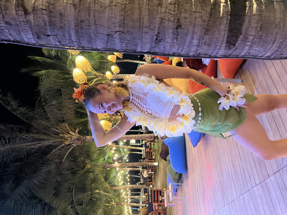 The image displays an outdoor nighttime scene located at a resort or lounge area in Nha Trang, Vietnam. A light-skinned female subject, exhibiting vitiligo on her face and arms, is positioned in the mid-ground, smiling and posing with her left arm raised, hand near her head. She wears a white crocheted halter-neck crop top, a yellow and white plumeria lei, and a green sarong secured by a cowrie shell belt. An orange flower is present in her hair, and she holds a small bouquet of plumeria flowers in her left hand. The background features multiple tall palm trees under a dark sky, illuminated by strings of warm-toned lights and decorative, spherical woven lamps. The ground consists of a light-colored wooden deck on which various colorful beanbag chairs (blue, green, orange, red, yellow) are scattered, along with small, low tables equipped with gold-colored lamps. In the far background, several indistinct figures are visible, seated at tables in a populated lounge area. A person in red clothing is partially visible behind an orange beanbag in the lower right foreground.