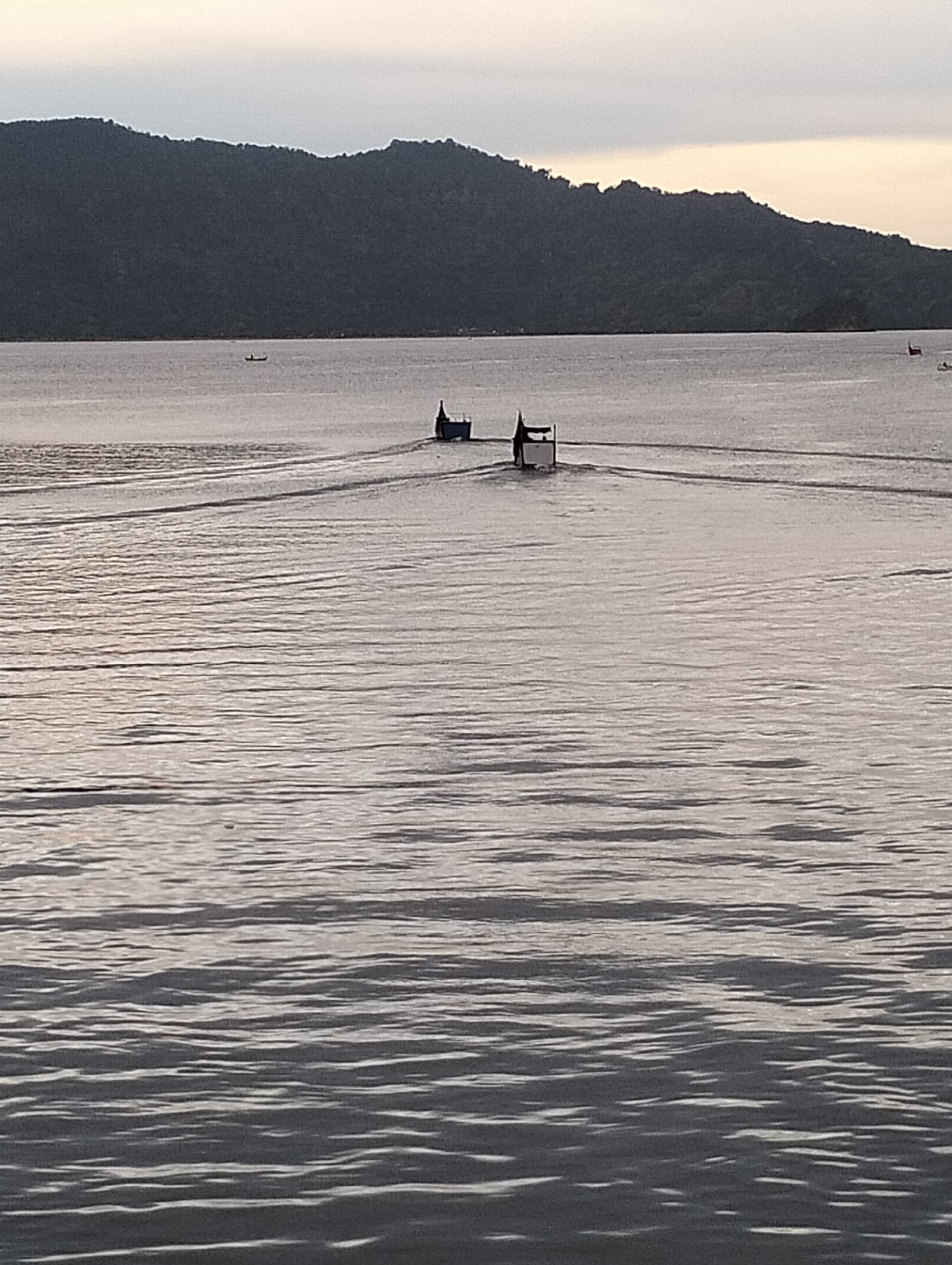 The photo shows two boats on a body of water. The water is relatively calm, with gentle ripples. A landmass is in the background.
One boat, in the middle distance, is traveling away from the viewpoint, leaving a wake in the water. A structure on the boat suggests a possible canopy. The presence of this boat indicates human activity on the water.
Another boat is further away, partially obscured by the background.
The location is likely near Banda Aceh, Indonesia, as the geographical features, and the presence of small fishing boats are characteristics of the area. The landscape consists of water, boats, and a landmass, which collectively illustrate the location in question. The people are likely locals who use the boats for fishing.